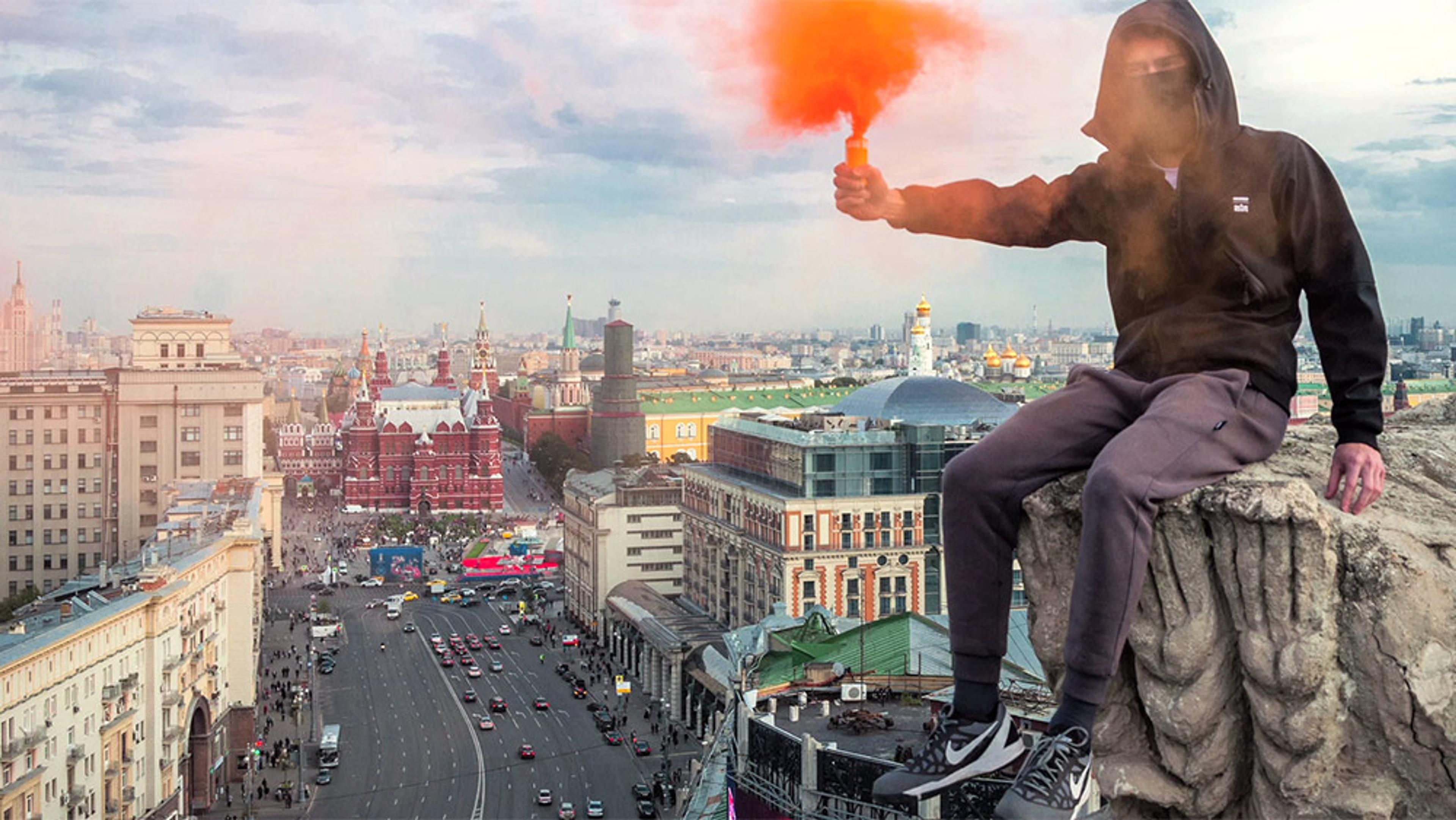 A young man in a hoodie and face mask holds a red smoke flare atop a high building overlooking a bustling Moscow cityscape.