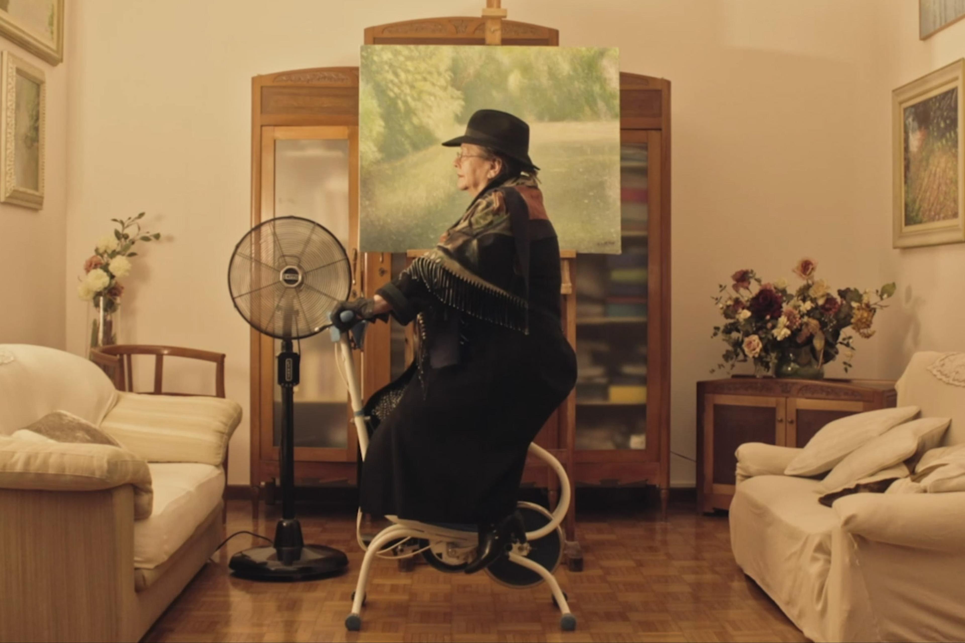 Older woman on a stationary bike in front of a painting near a fan in a living room. There are flowers and sofas in the background.