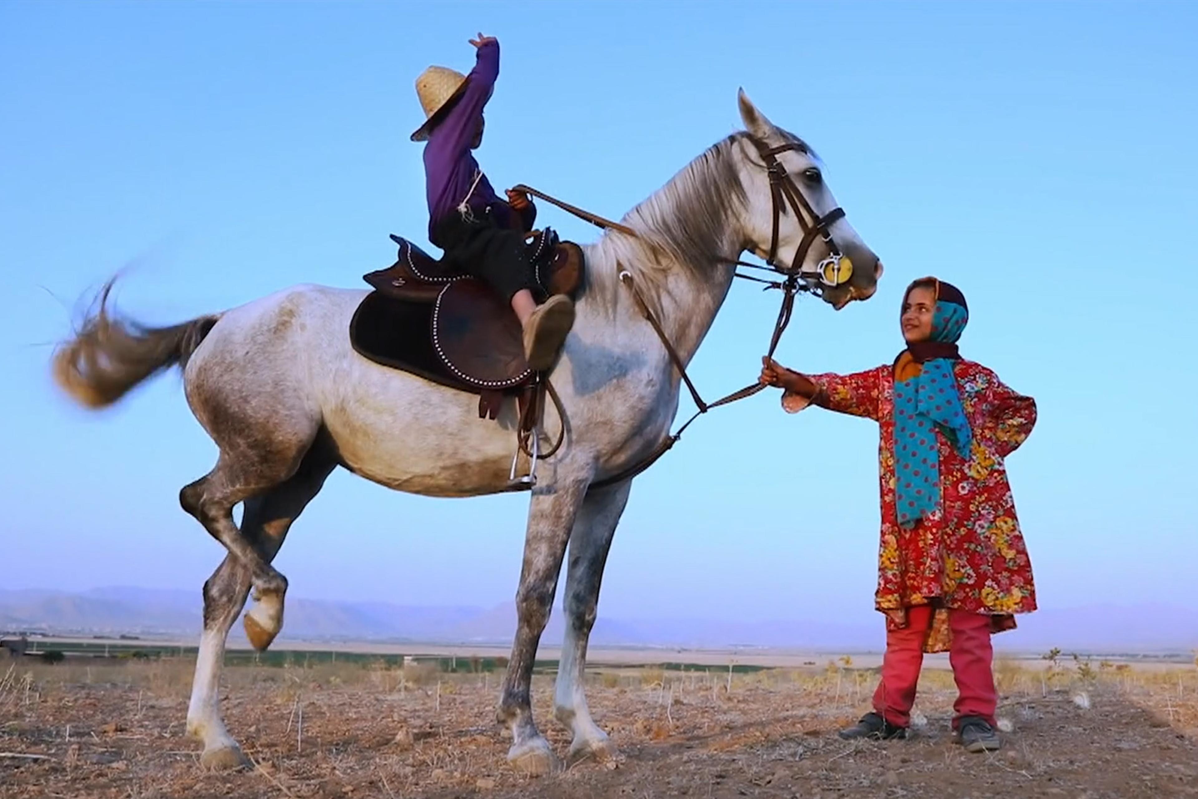 Photo of a boy riding a rearing horse while a girl in colourful clothing holds the reins, with a clear sky and dry landscape.