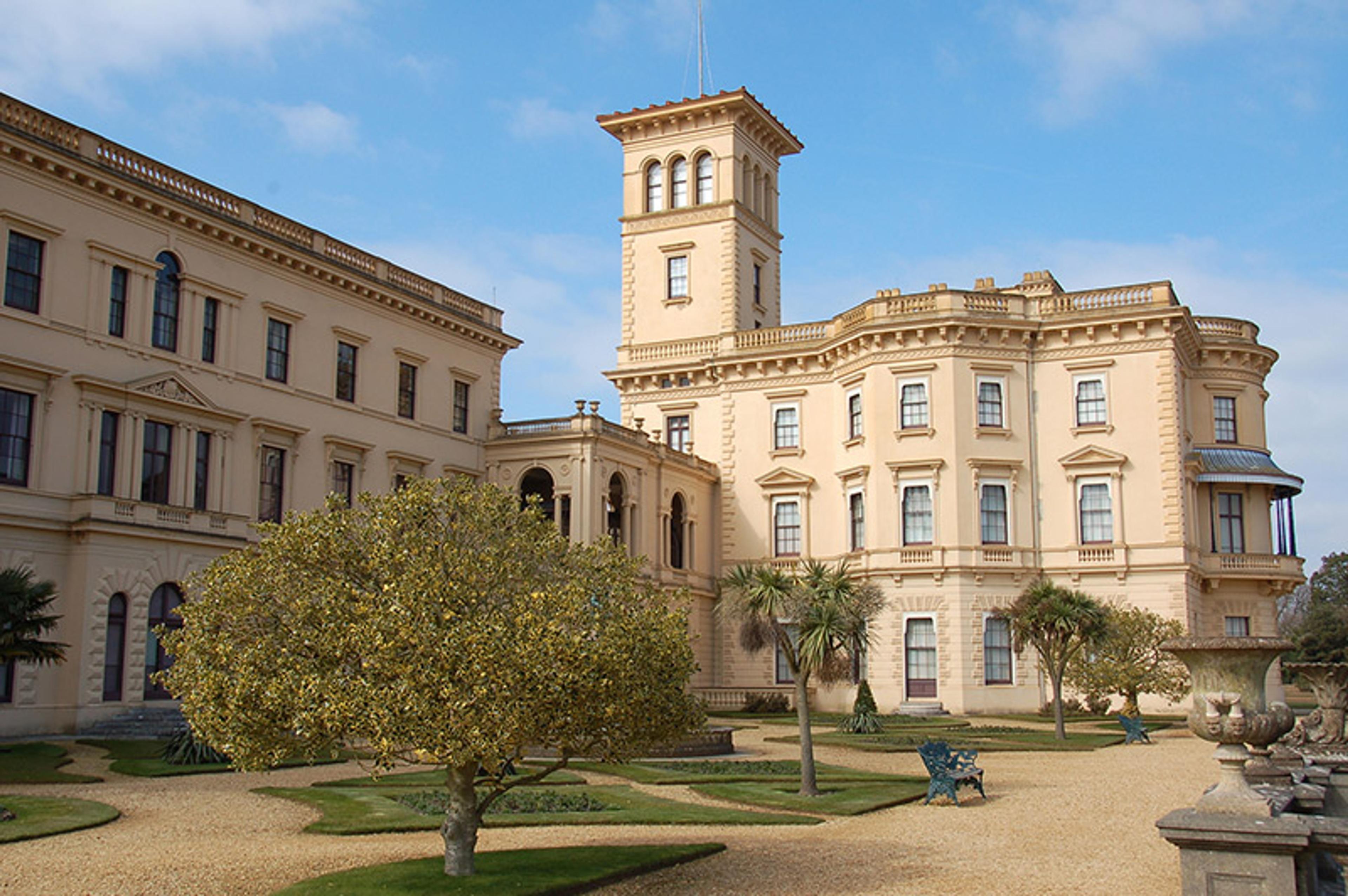 A grand, historic building with beige stone walls, large windows, a central tower, and a landscaped garden with trees.