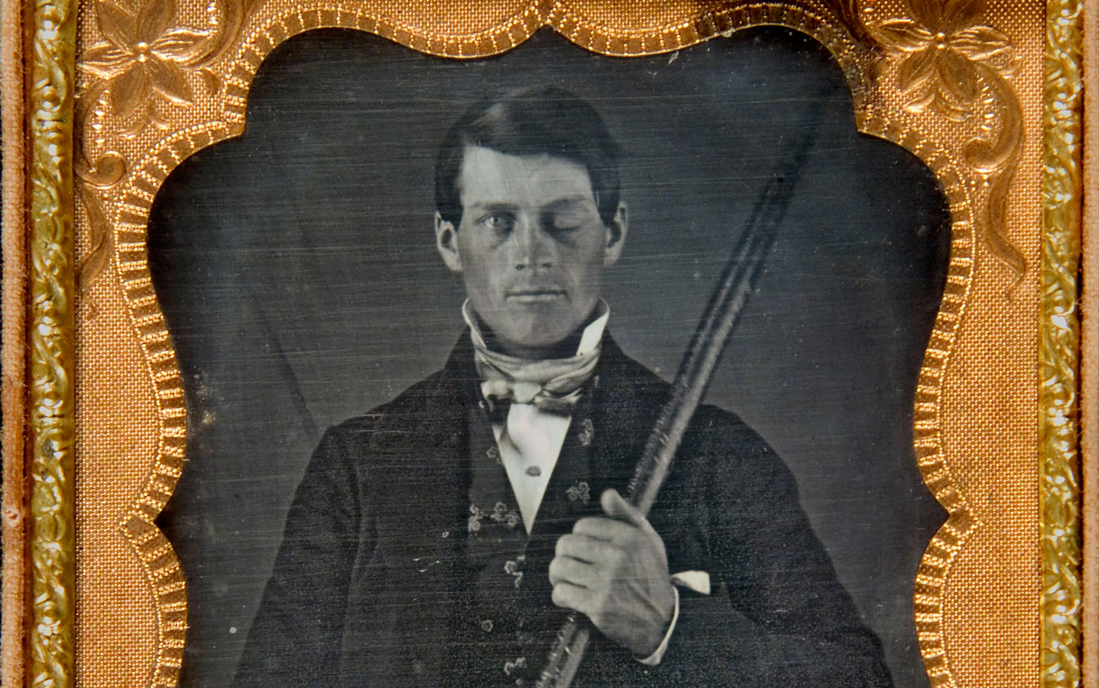 A black-and-white daguerreotype of a man in a formal suit holding a rod, one eye appears damaged, framed in an ornate gold border.