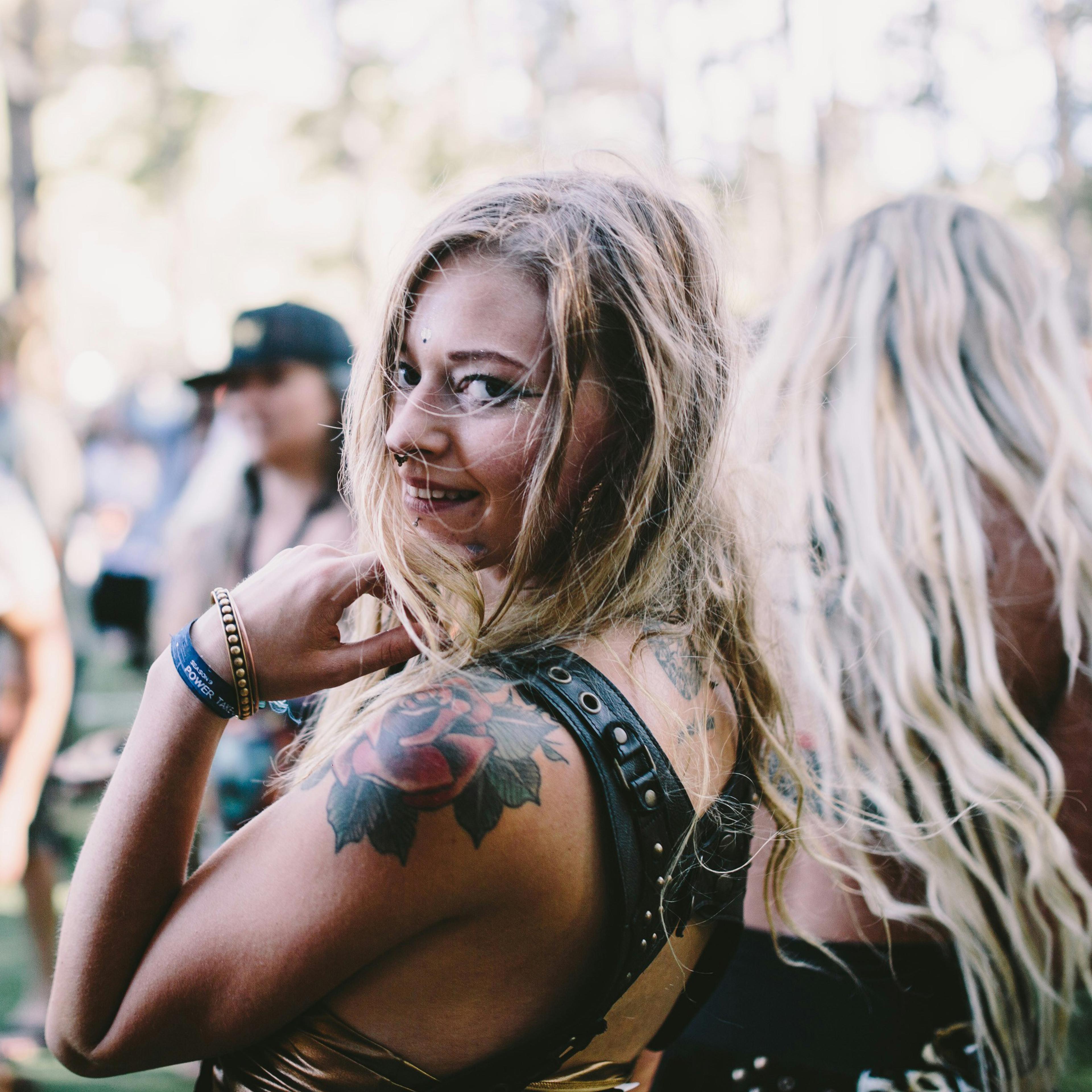 A smiling woman with tattoos at an outdoor festival, surrounded by blurred people and trees in the background.