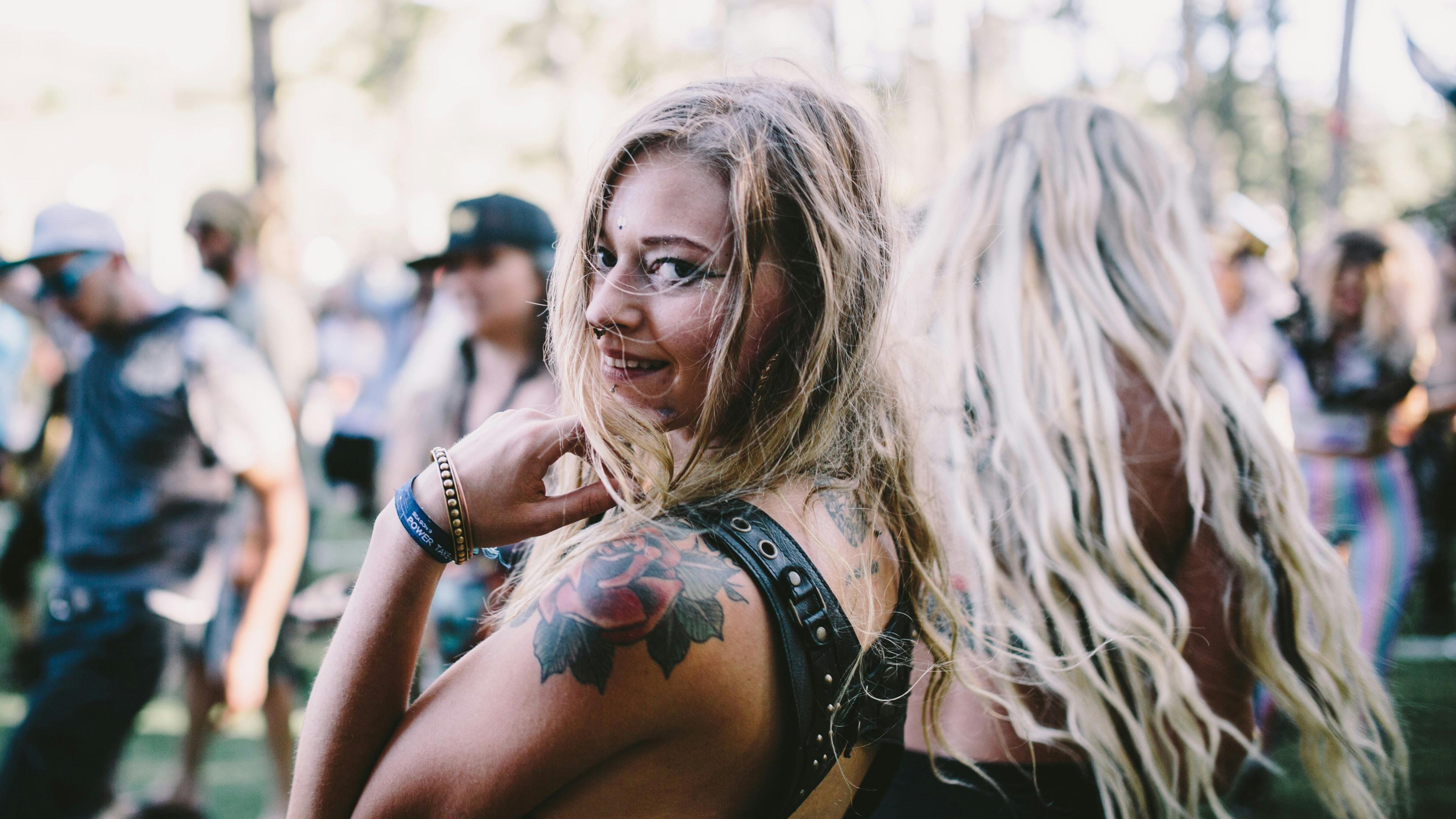 A smiling woman with tattoos at an outdoor festival, surrounded by blurred people and trees in the background.