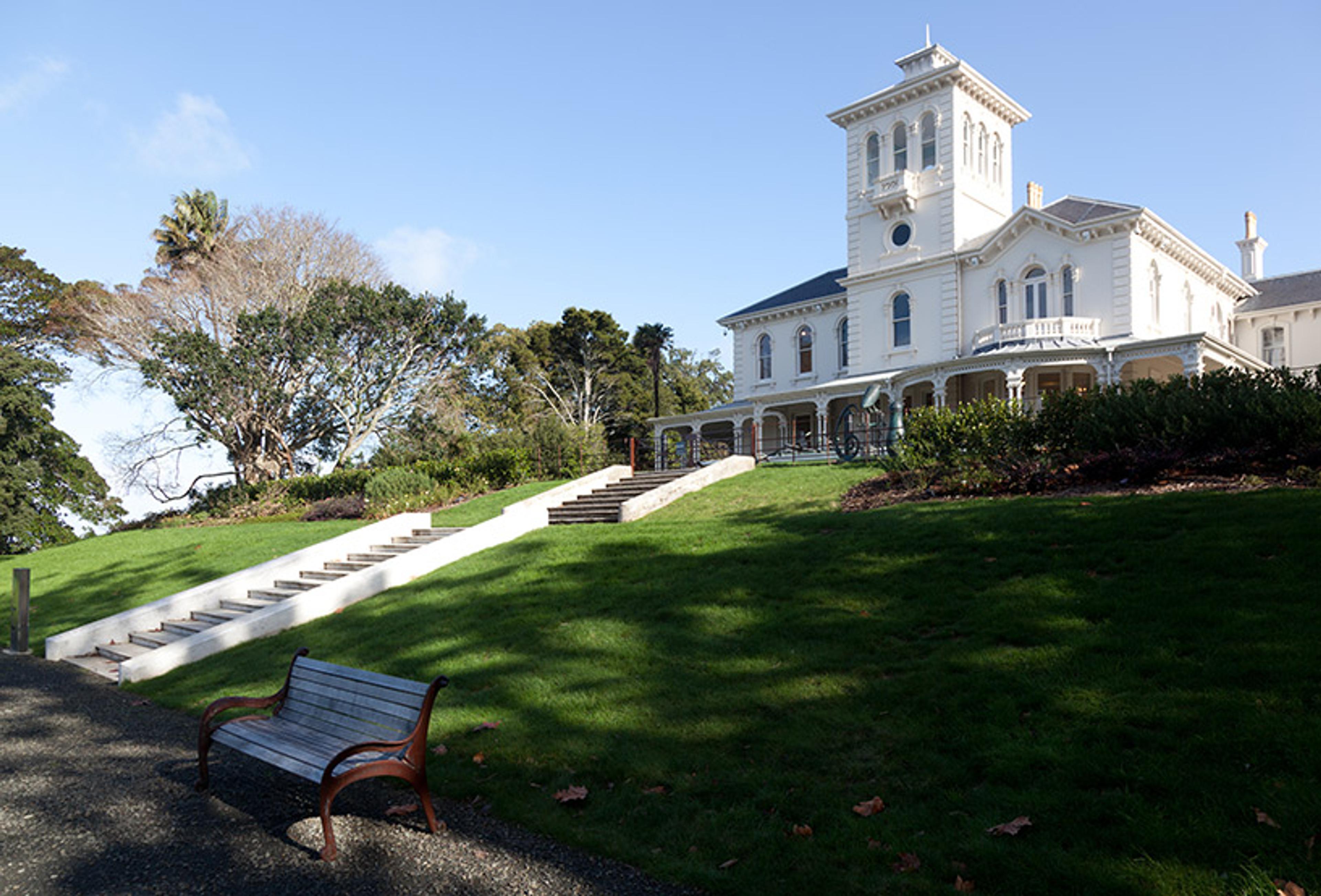 A large white historic building with a tower, surrounded by trees and greenery, seen from the base of a hill with a bench and steps.