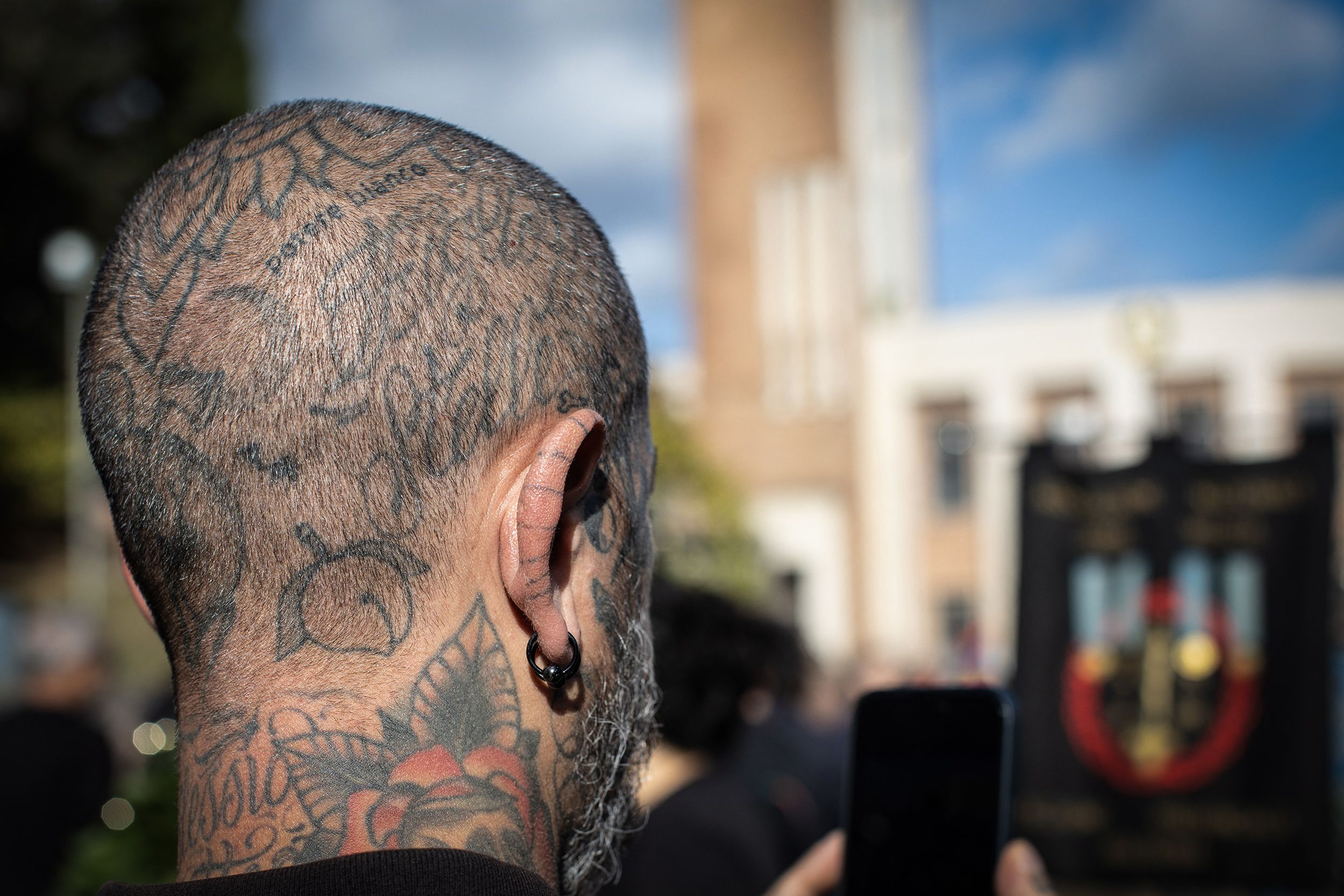 Close-up of a man with tattooed scalp and piercings, holding a phone in a crowd, with a building and banner in the background.