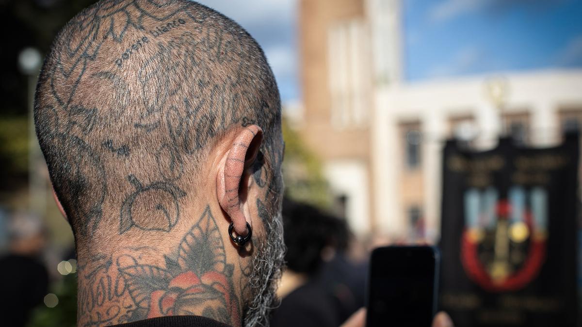 Close-up of a man with tattooed scalp and piercings, holding a phone in a crowd, with a building and banner in the background.