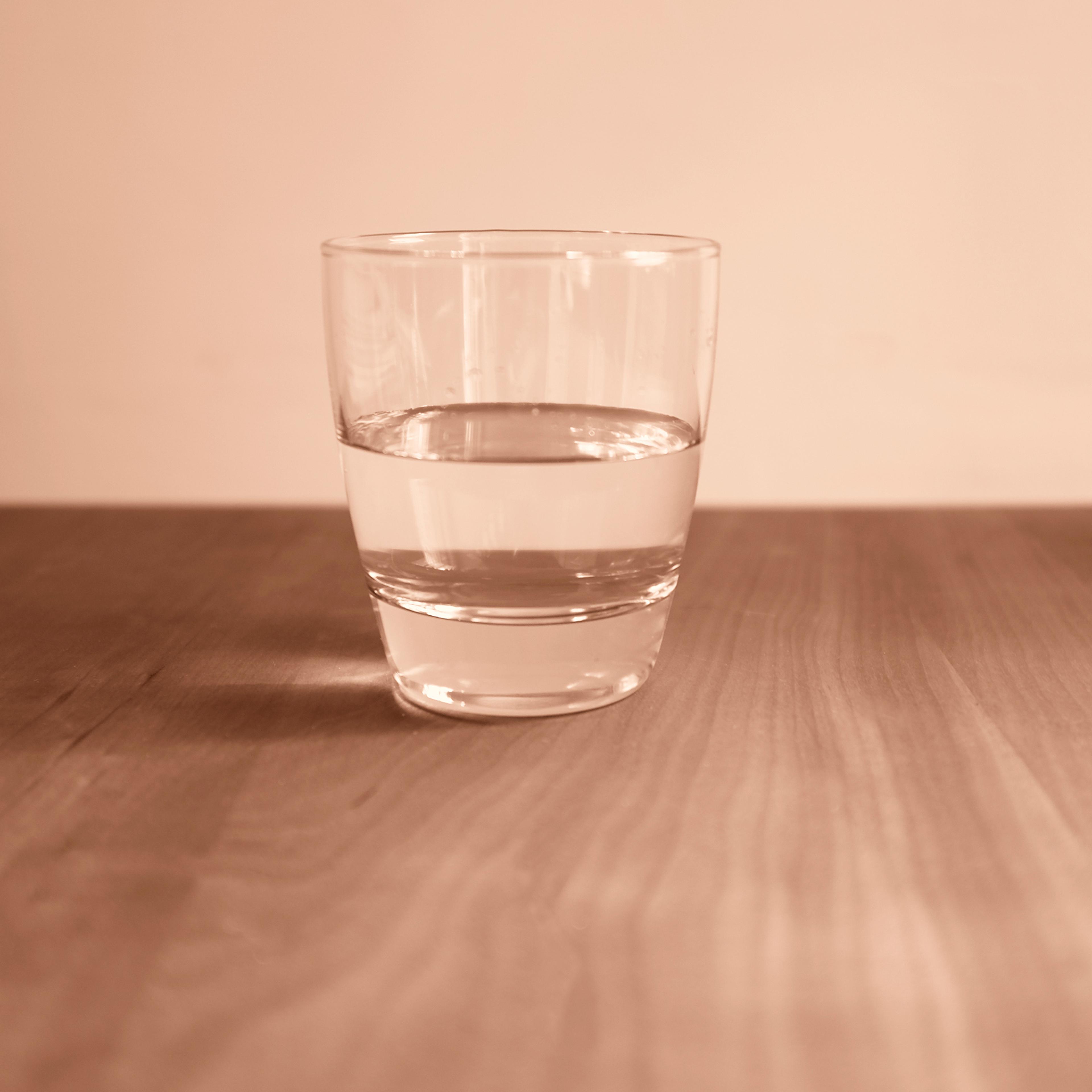 A clear glass, half-filled with water, placed on a wooden surface. The background is a plain, soft beige. The image has a warm, sepia tone.