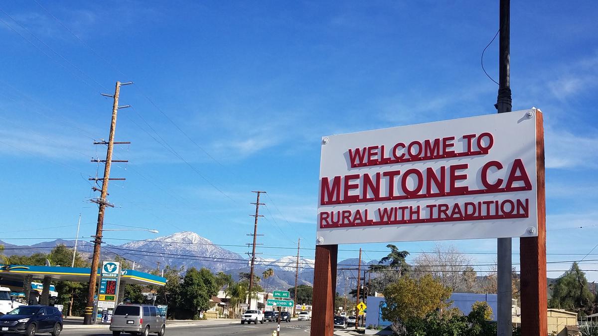 Photo of the Welcome to Mentone, CA sign with the tagline “Rural With Tradition”, against a mountain backdrop, clear blue sky above.