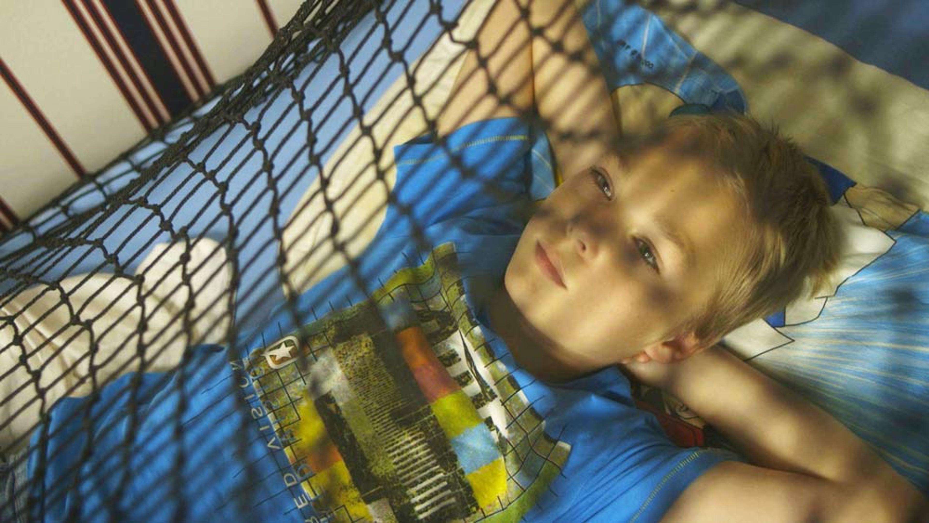 Photo of a boy in a blue shirt lying in a hammock with his arms behind his head, looking thoughtful. Shadow of net visible.