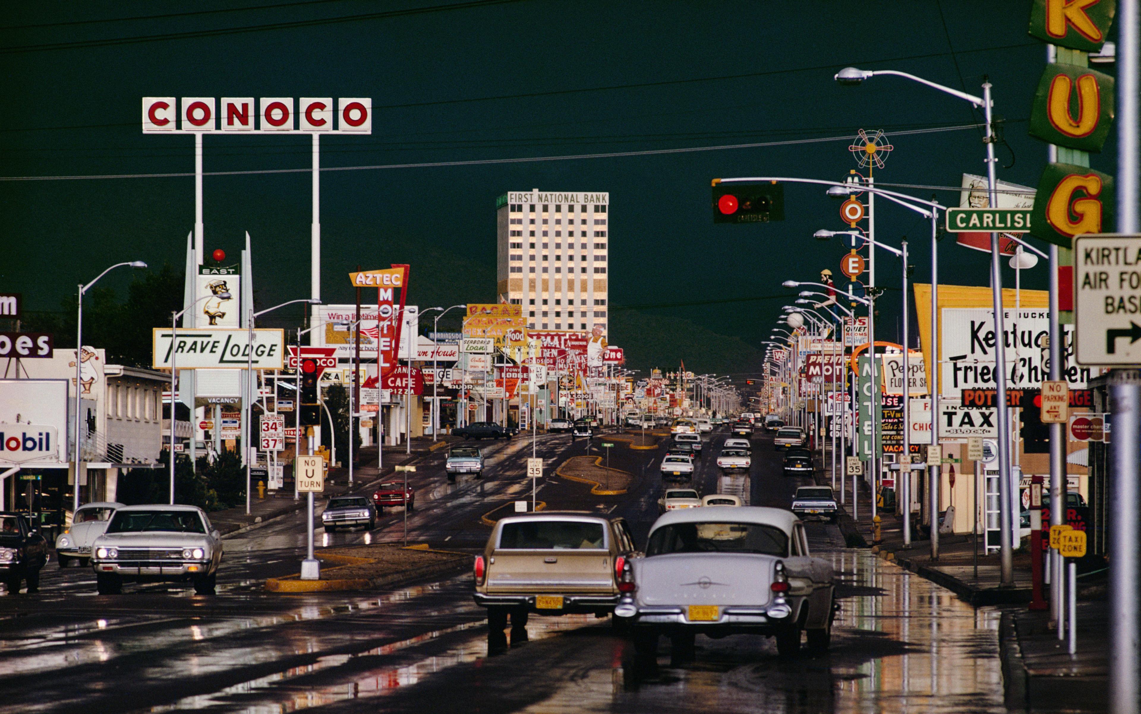 A busy city street at night with illuminated signs, cars on wet roads, and a tall building in the background.