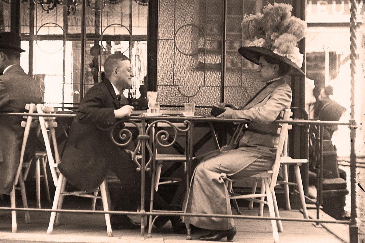 Vintage photo of a man and a woman in a large hat talking at an outdoor café, seated at a round table with drinks.