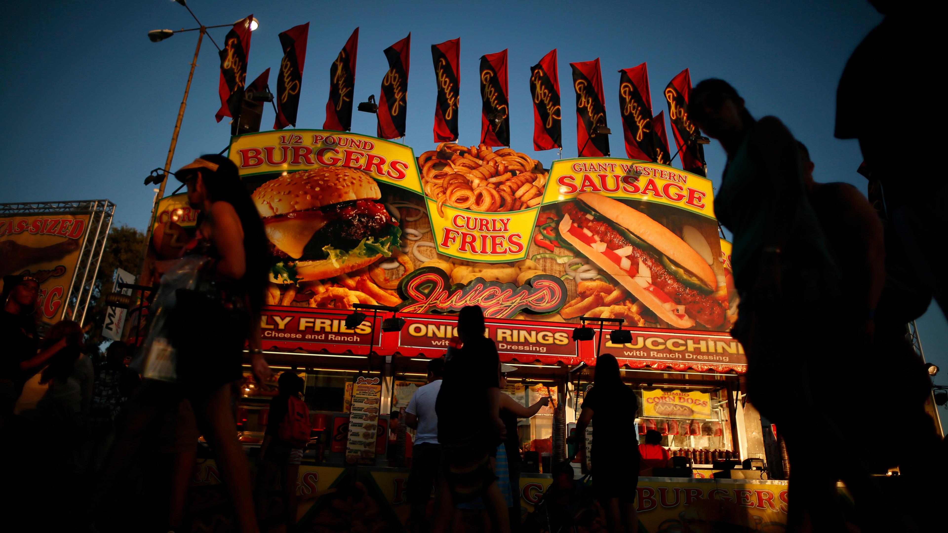 A bustling food stall at dusk with bright signs advertising burgers, curly fries and sausages against a darkening sky.