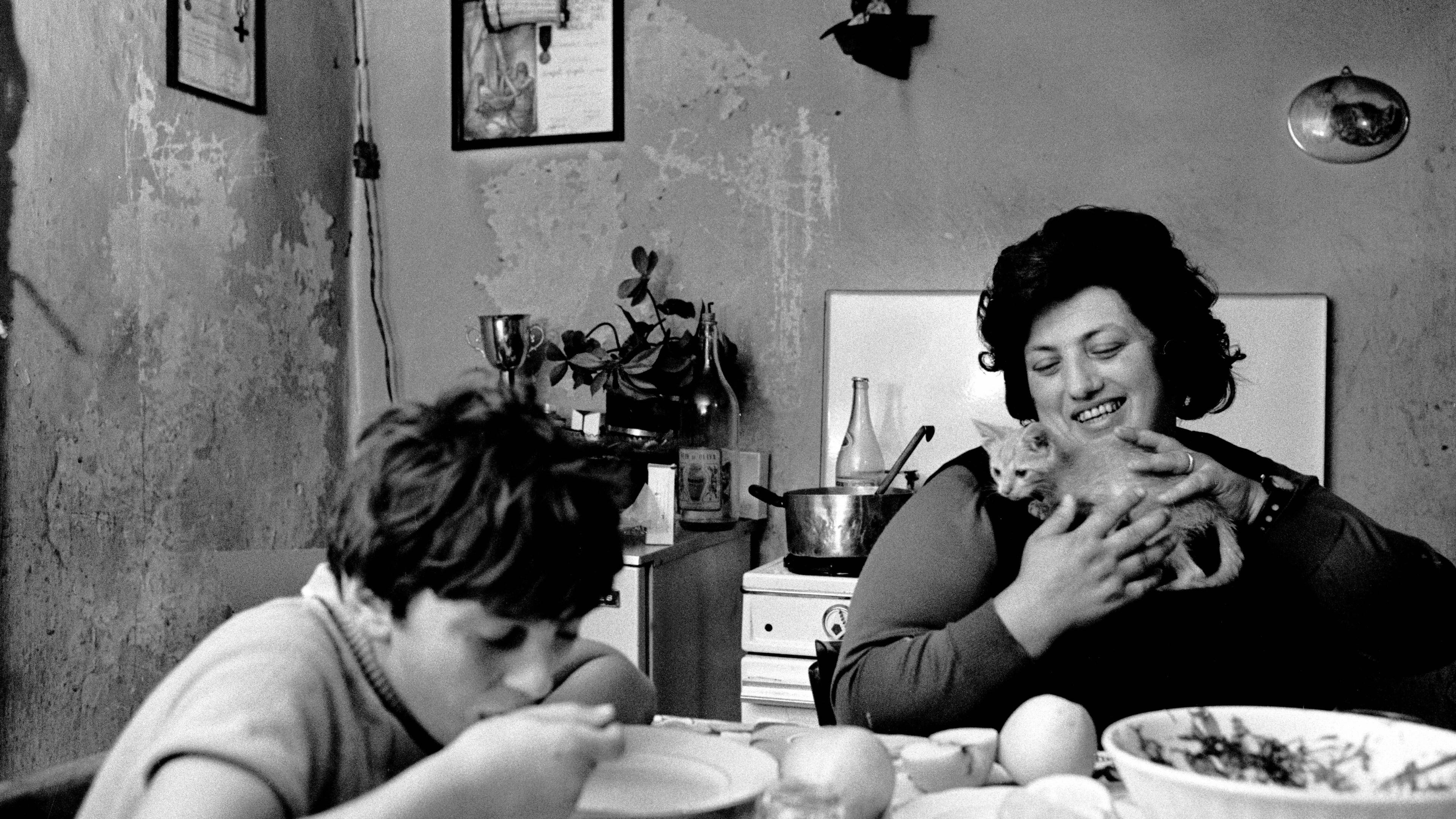 Black and white photo of a woman holding a cat in a kitchen while a child eats at the table.