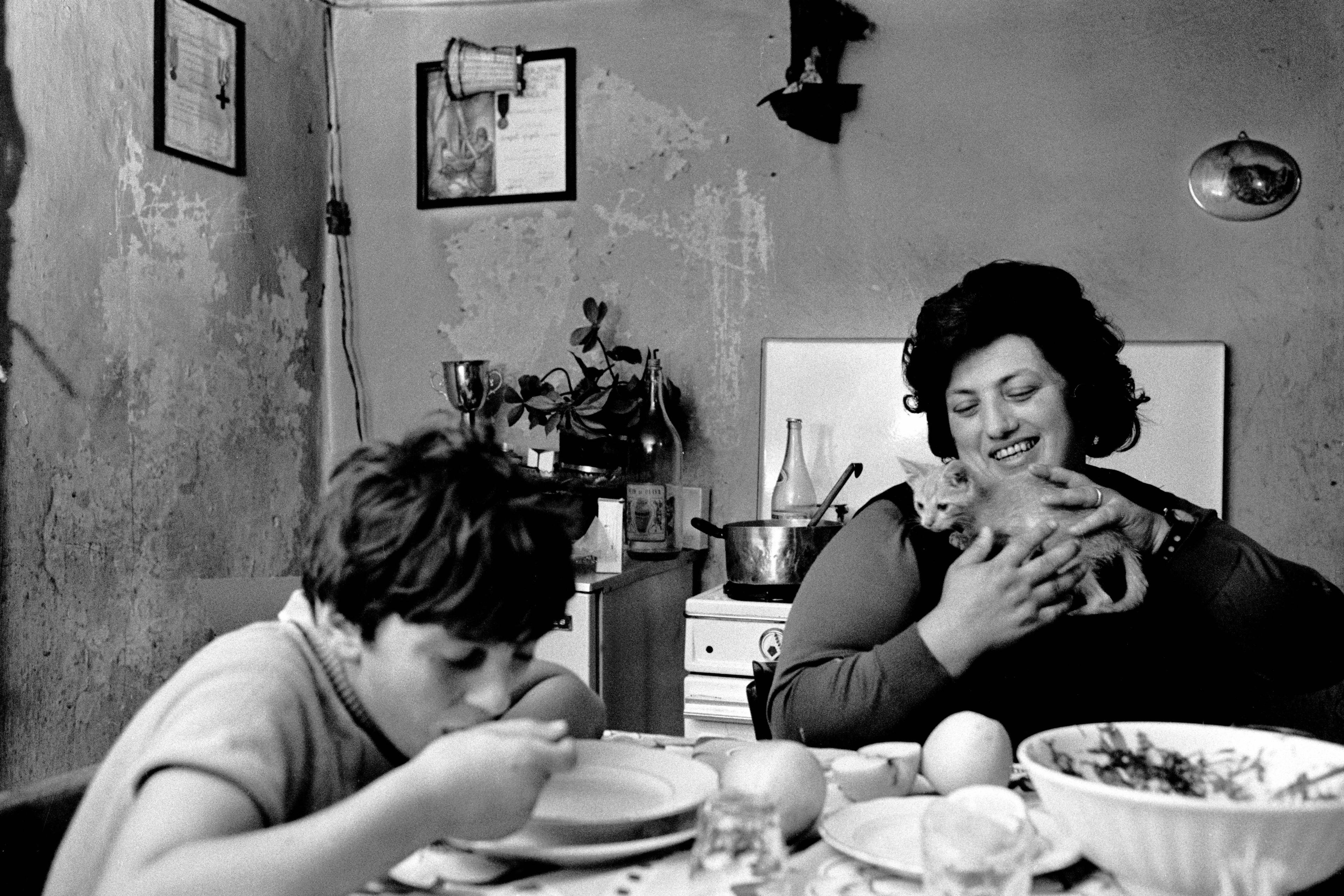 Black and white photo of a woman holding a cat in a kitchen while a child eats at the table.