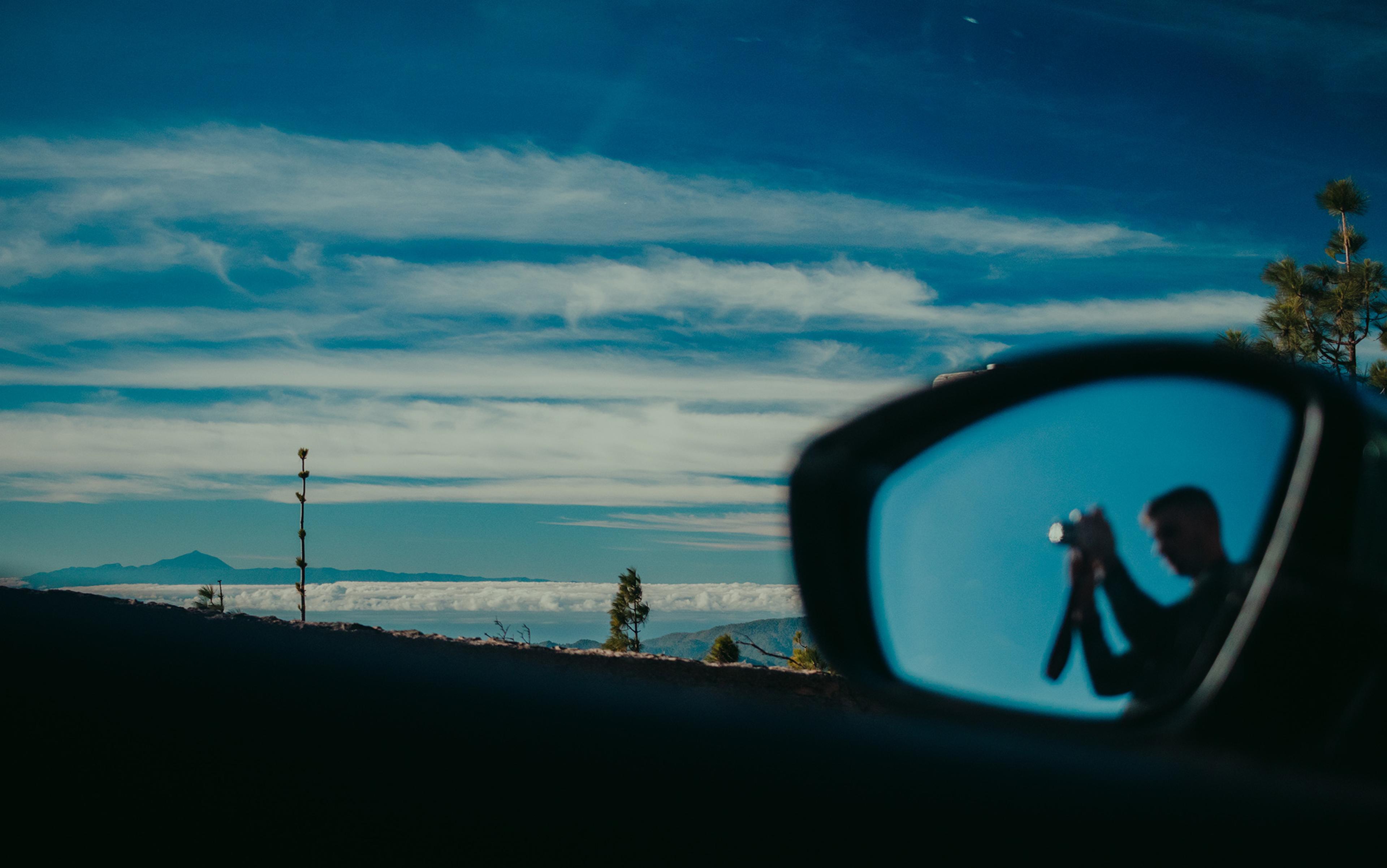 A landscape with blue sky and clouds, reflected in a car’s side mirror is a person holding a camera.