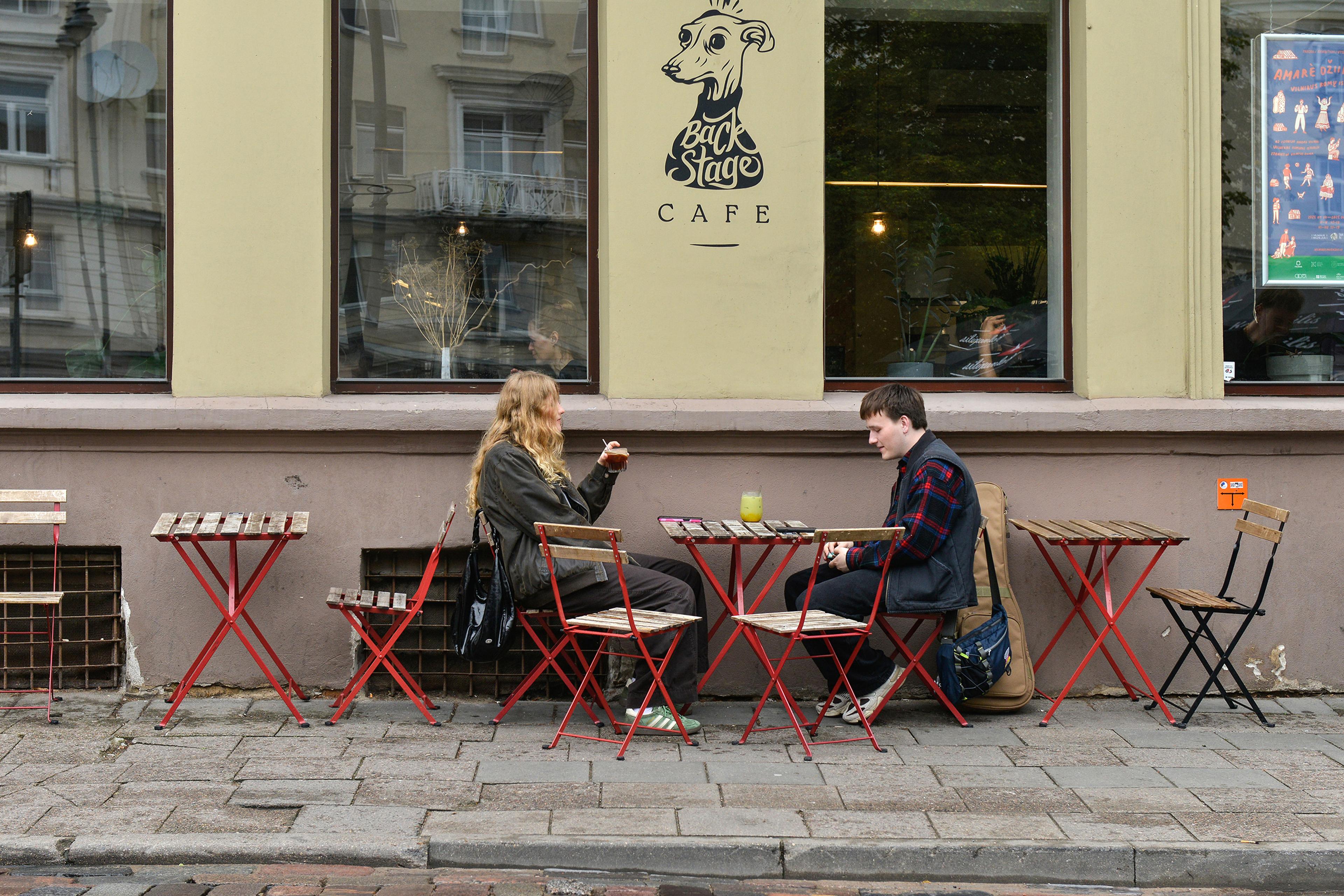 Two people seated at an outdoor café with red tables and chairs, talking in front of a large window.