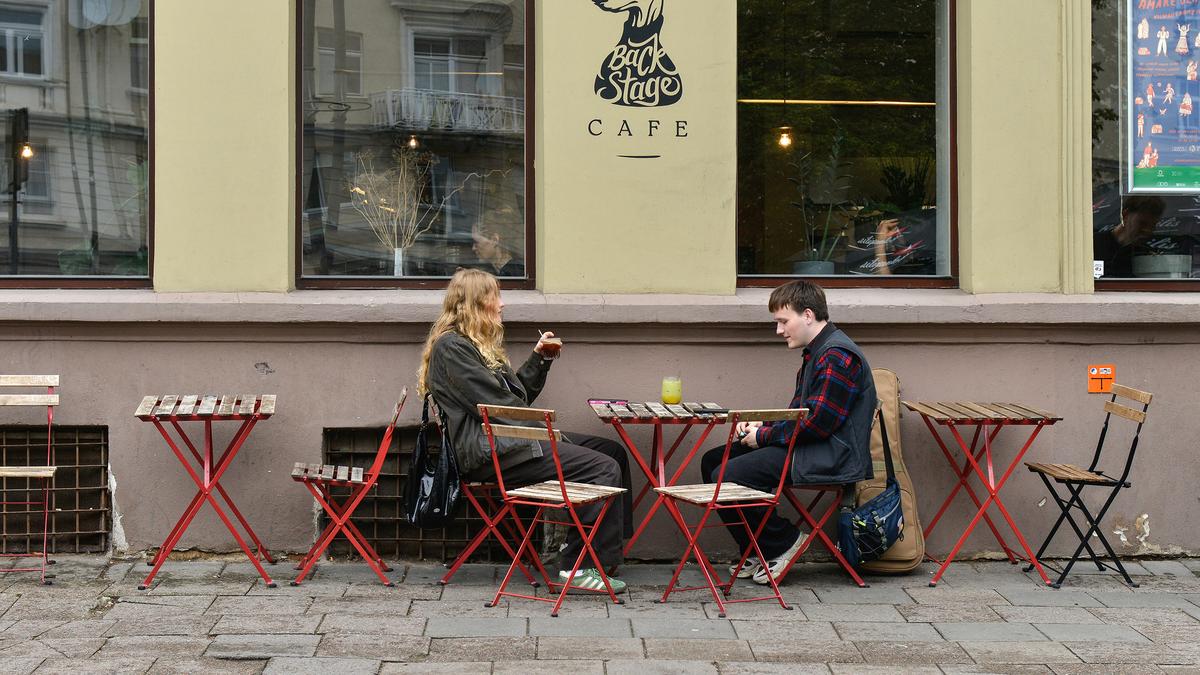 Two people seated at an outdoor café with red tables and chairs, talking in front of a large window.