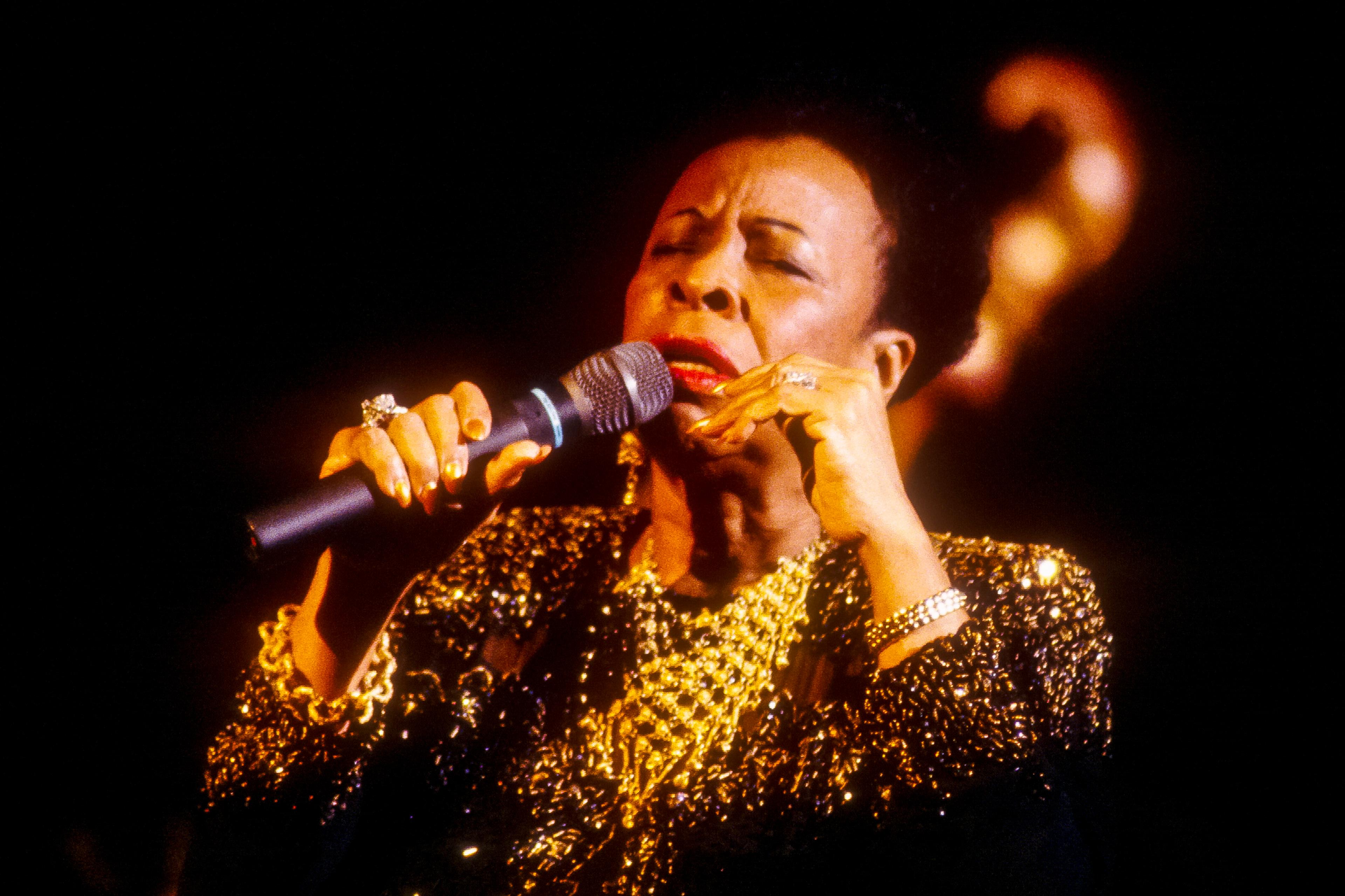 Photo of a woman singing passionately into a microphone wearing a sparkling black and gold dress on a dark stage.