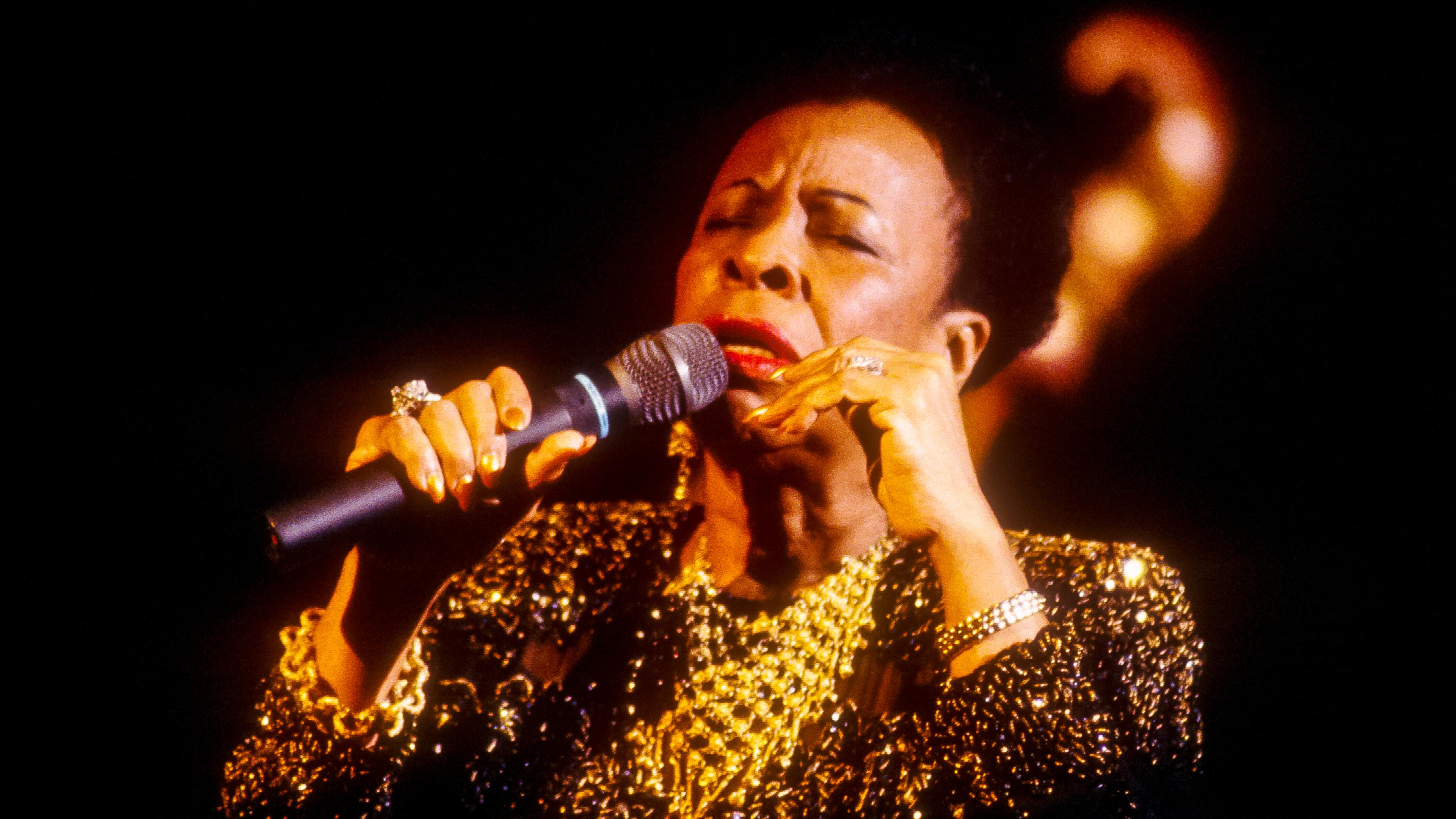 Photo of a woman singing passionately into a microphone wearing a sparkling black and gold dress on a dark stage.