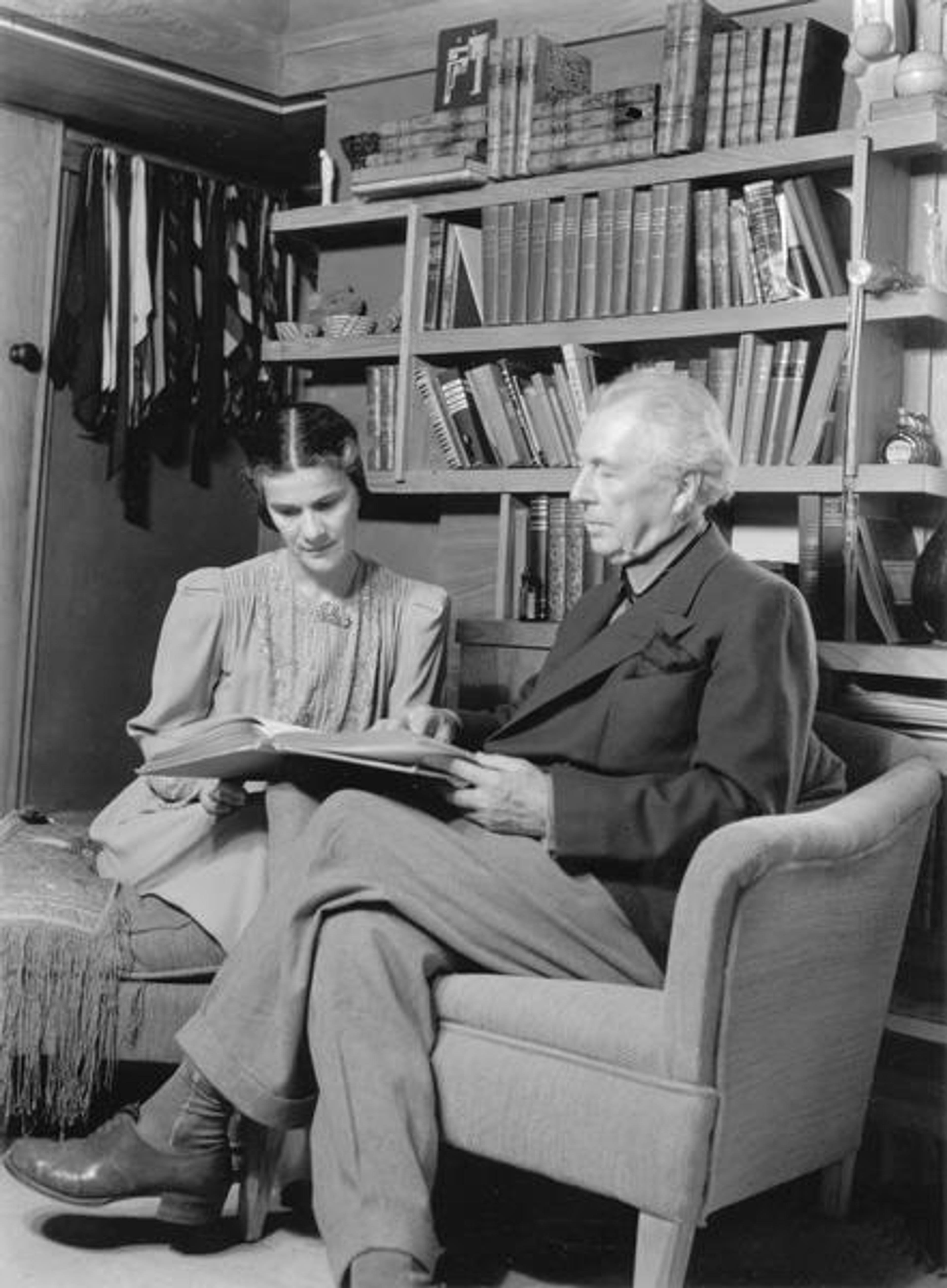 Black and white photo of a man and woman reading a book together in a room with bookshelves.