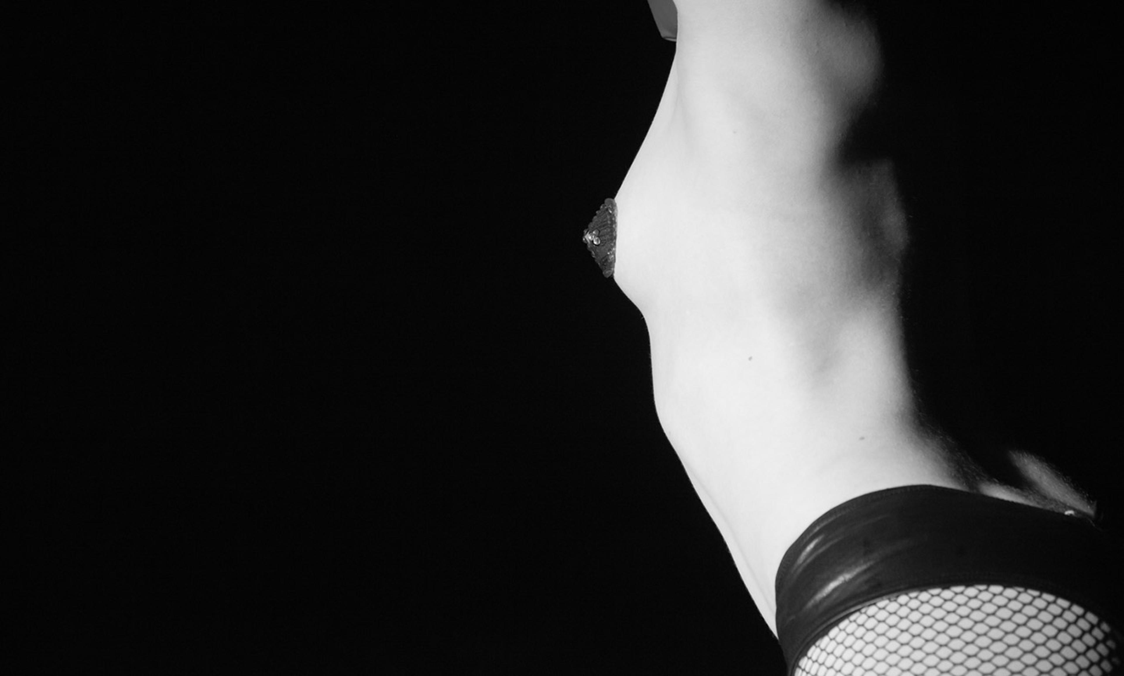 Black and white photo of a nude woman from the side showing their torso and thigh, partially clothed in fishnet stockings against a dark background.