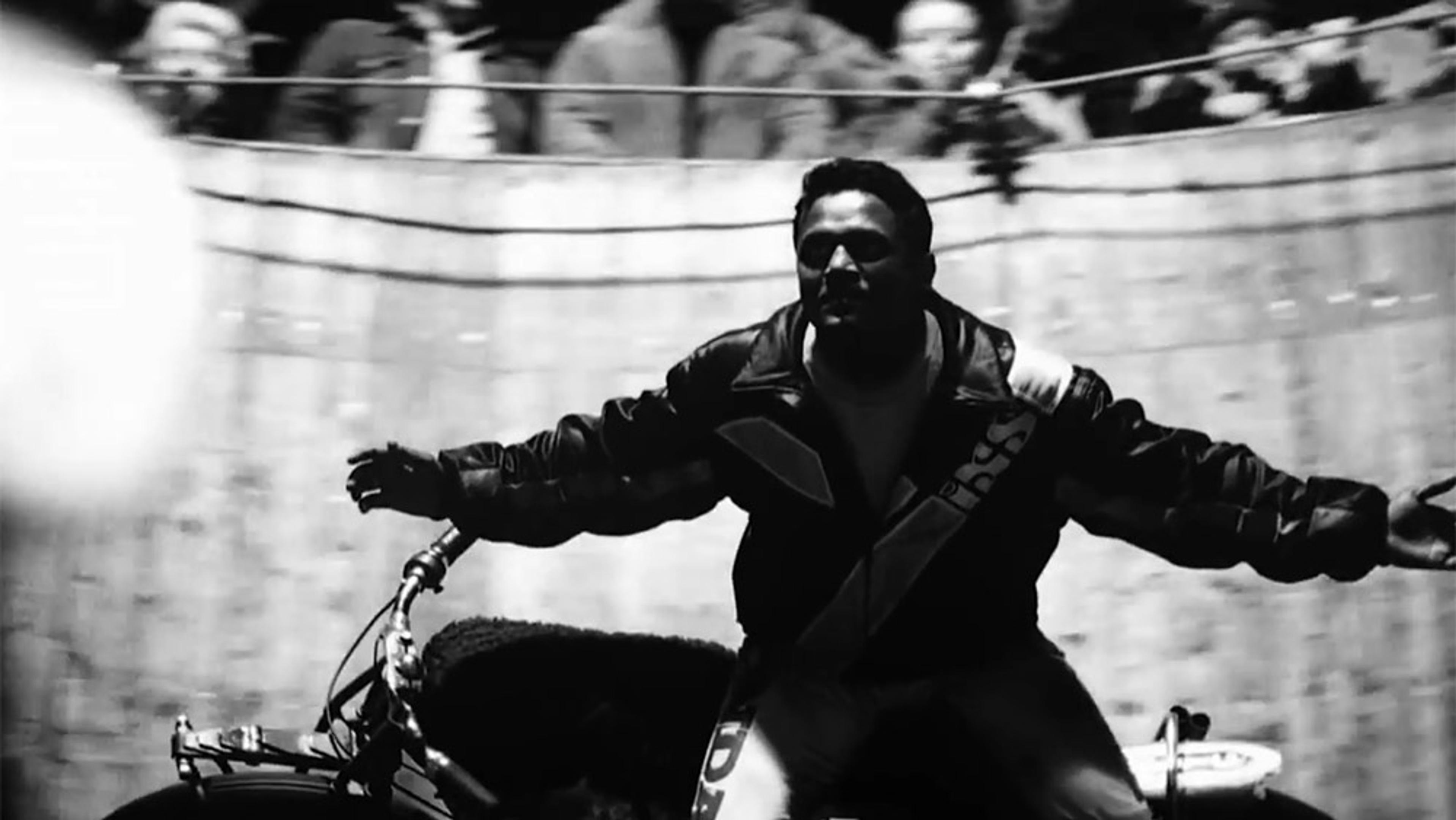 Black-and-white photo of a man with outstretched arms riding a motorcycle inside a wooden wall of death with spectators above.