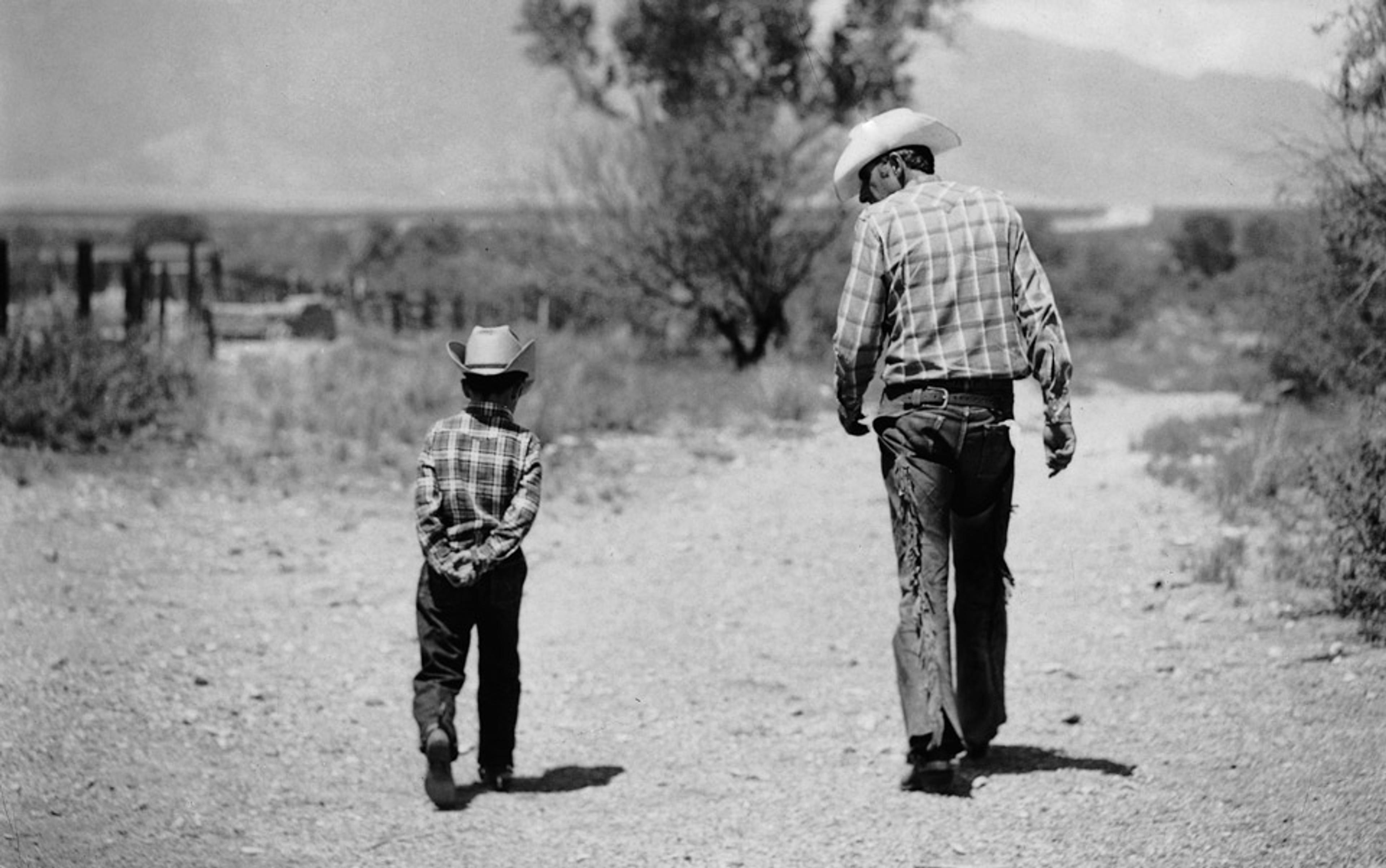 Black-and-white photo of a man and a boy wearing cowboy hats and flannel shirts, walking away on a dirt path in a rural setting.