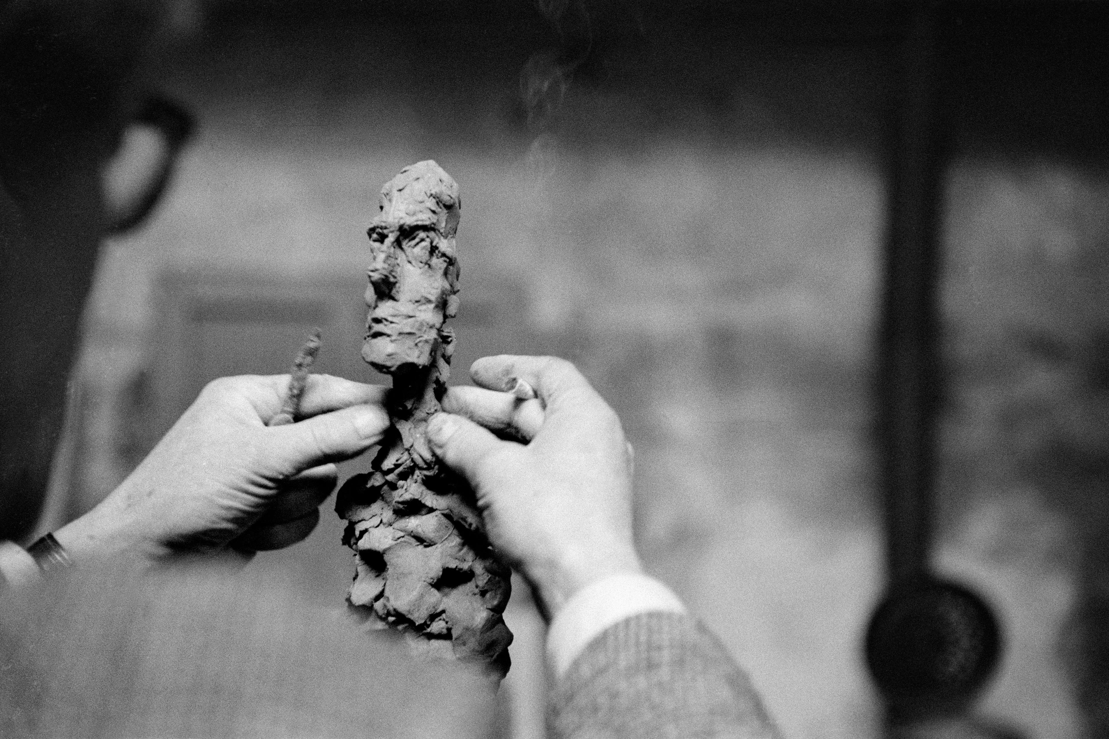 Black-and-white photo of hands sculpting a small clay figure with a rough-textured face in an art studio setting.