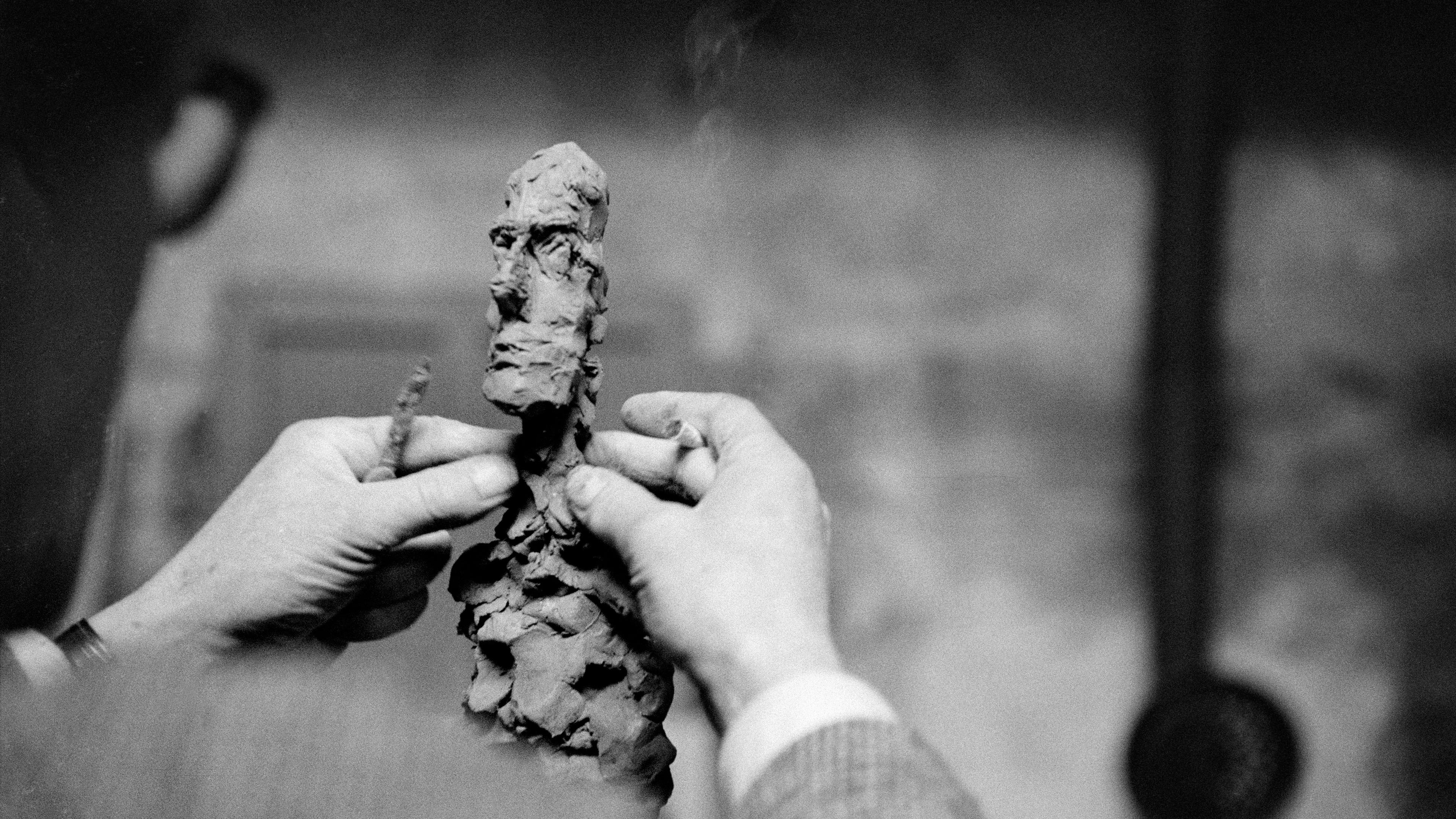 Black-and-white photo of hands sculpting a small clay figure with a rough-textured face in an art studio setting.