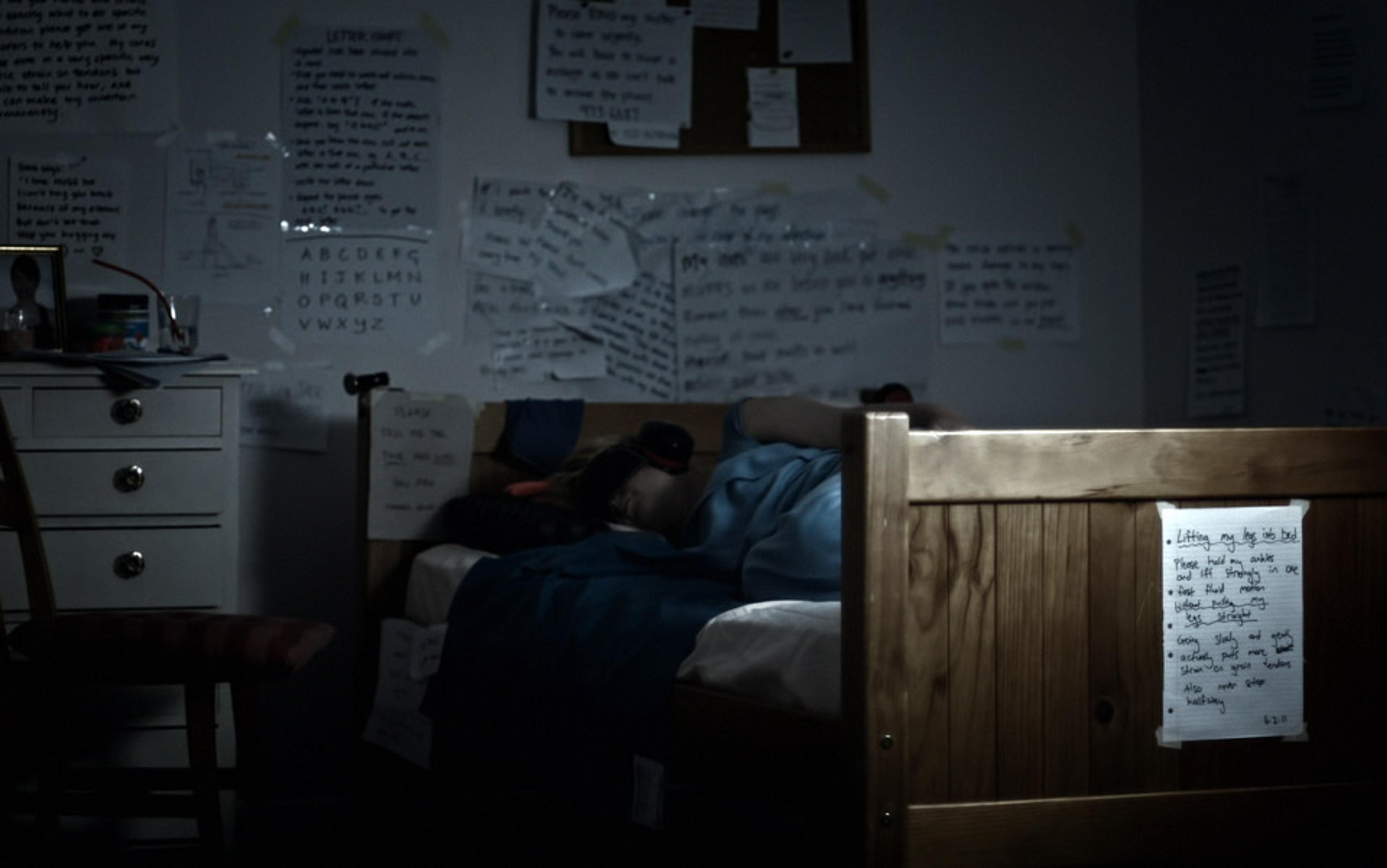 A dimly lit bedroom. A person lies in bed with notes and papers covering the walls, dresser, and footboard.