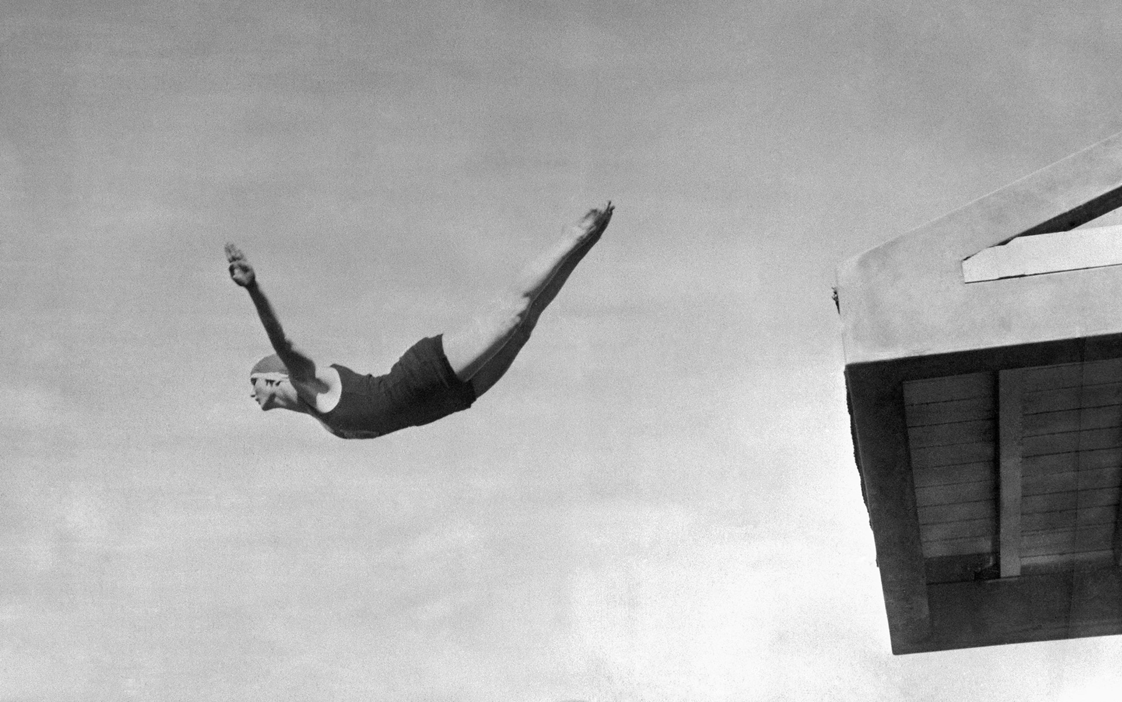 Vintage photo of a female diver in mid-air near a diving board, arms extended, body taut, in flight against a clear sky.