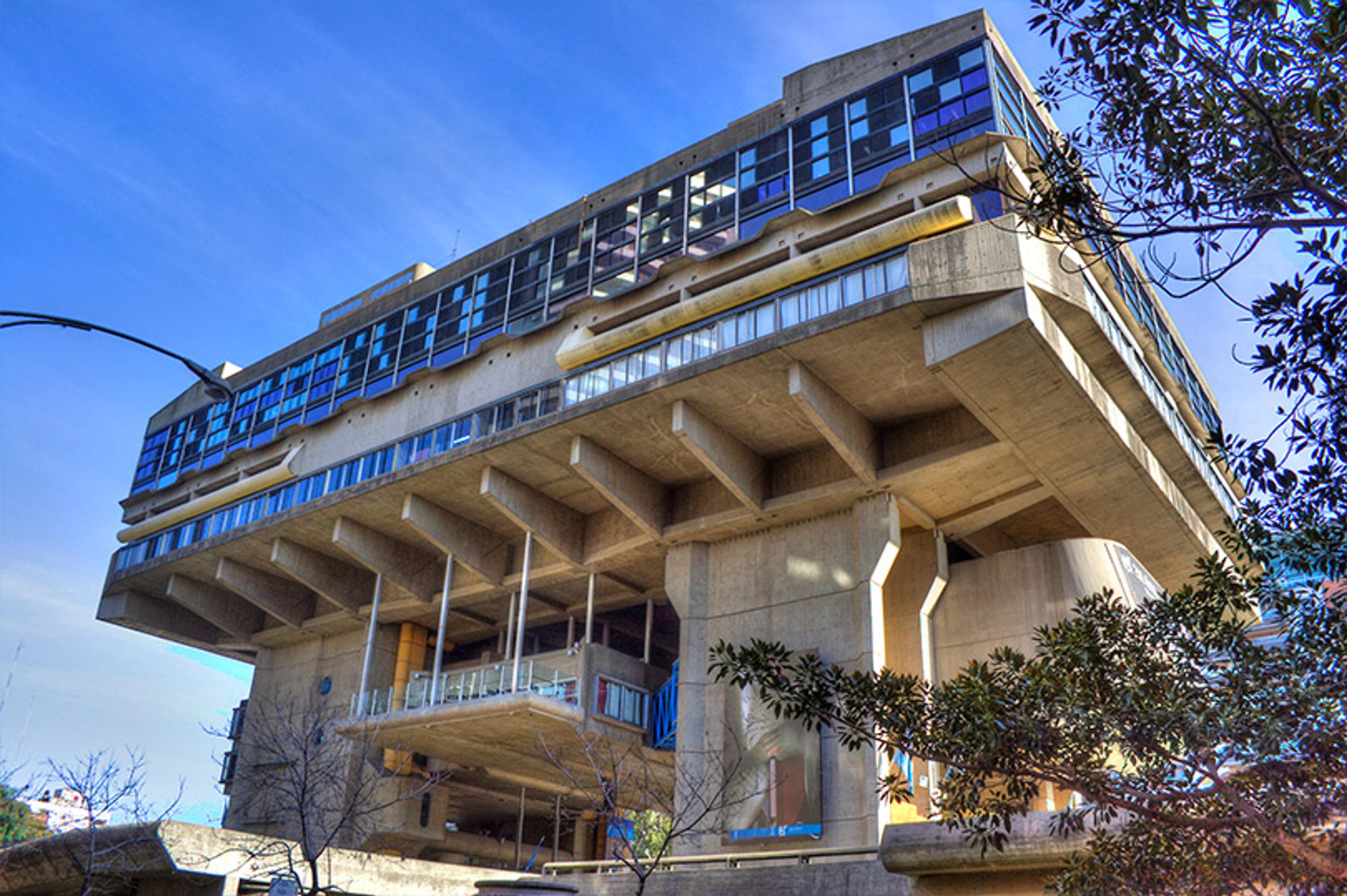A modern concrete building with bold, squared lines elevated on pillars, surrounded by trees against a blue sky.