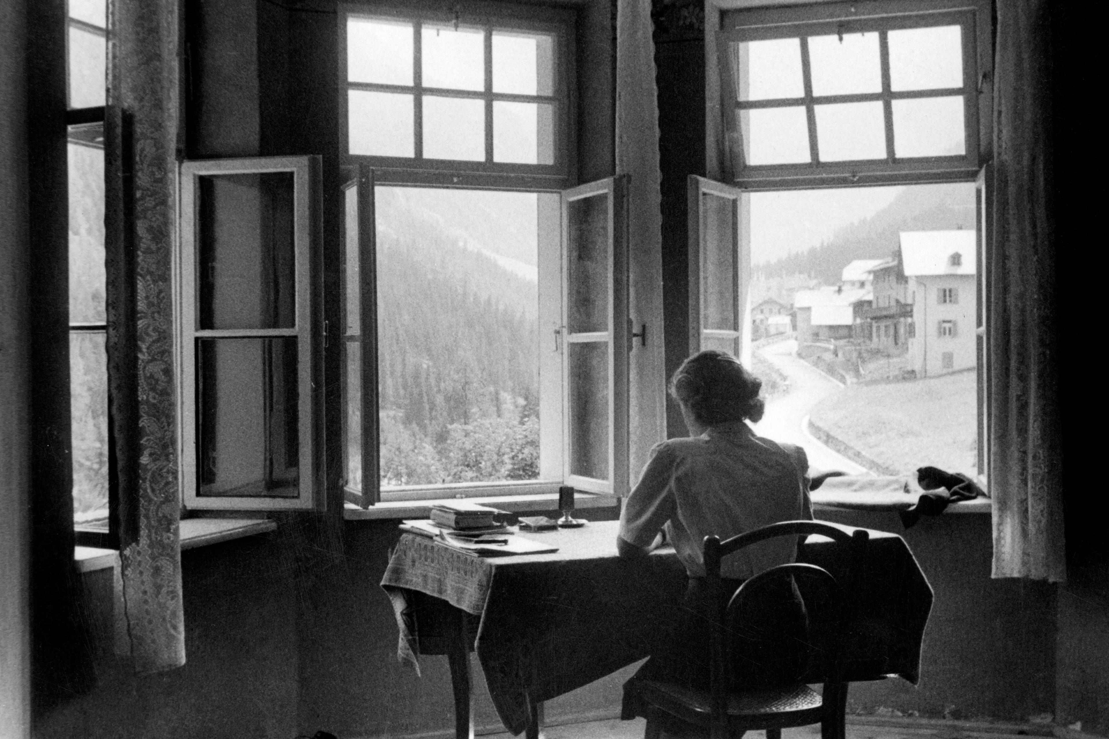 Black and white photo of a person sitting at a table in front of open windows with a mountain village view.