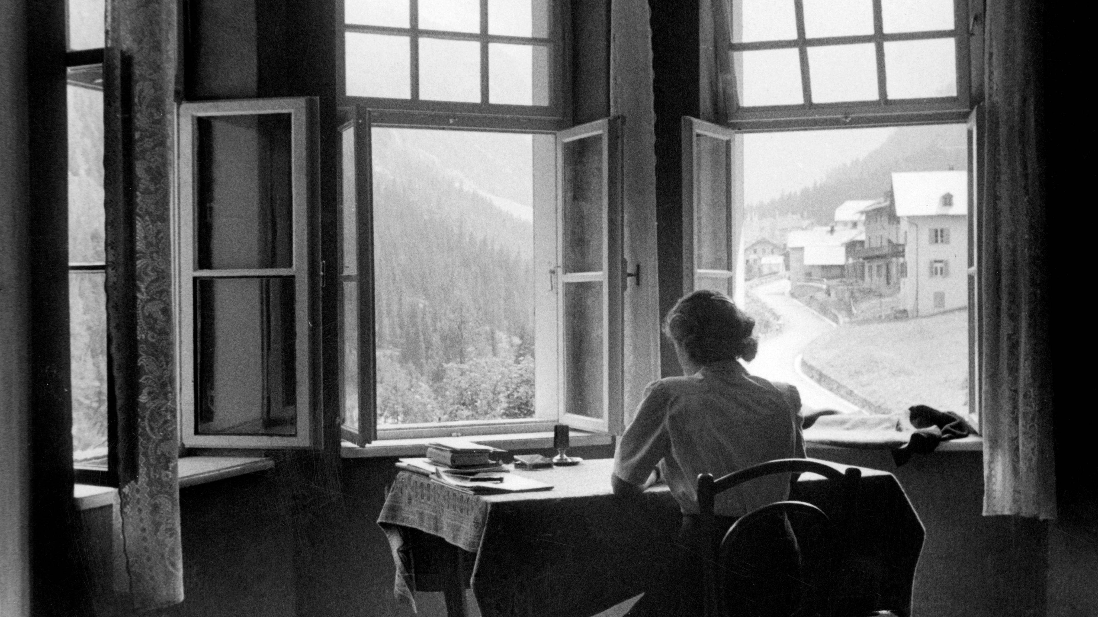 Black and white photo of a person sitting at a table in front of open windows with a mountain village view.