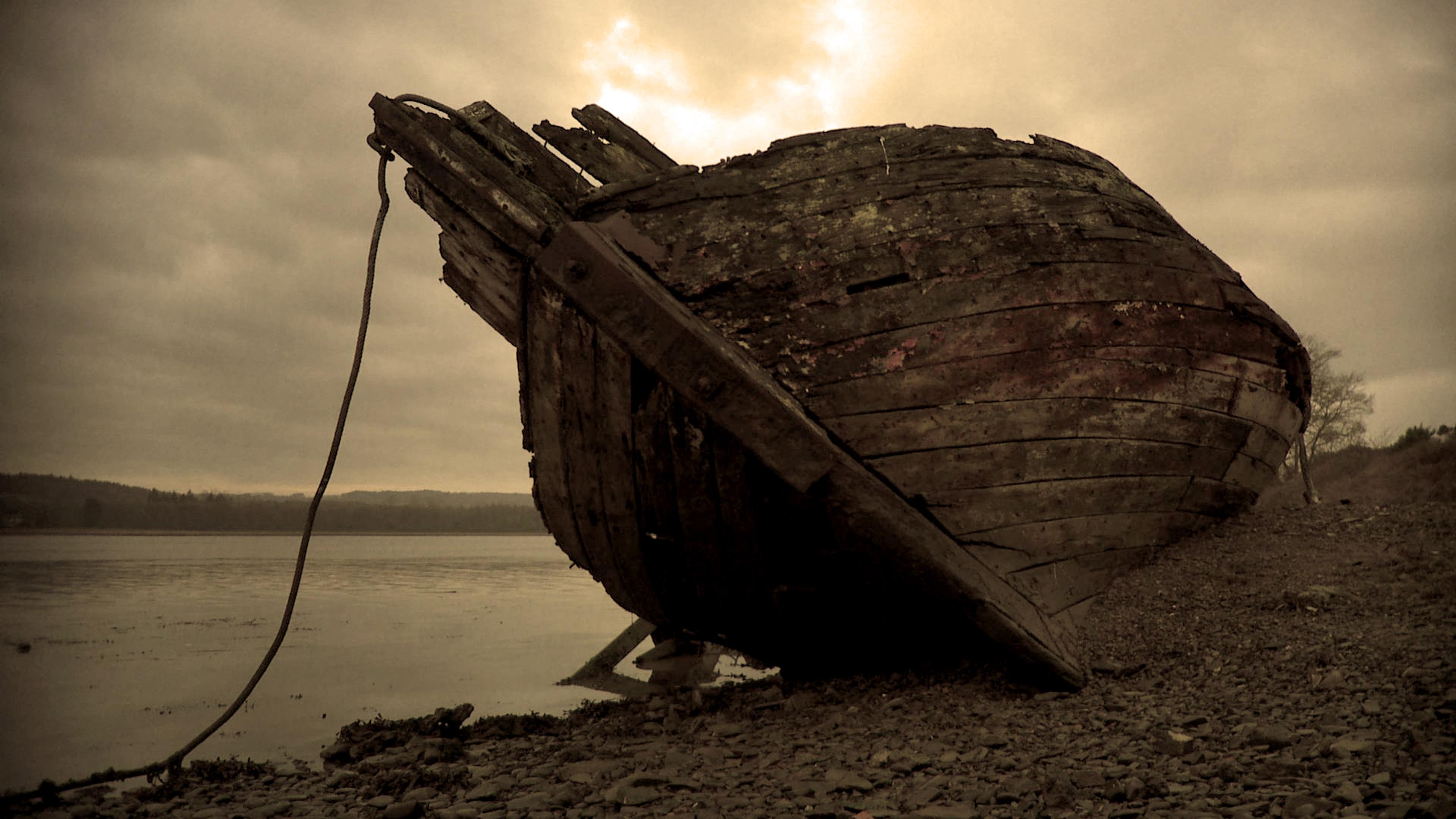 An old wooden boat decaying on a rocky shore by a calm lake with a cloudy grey sky in the background.