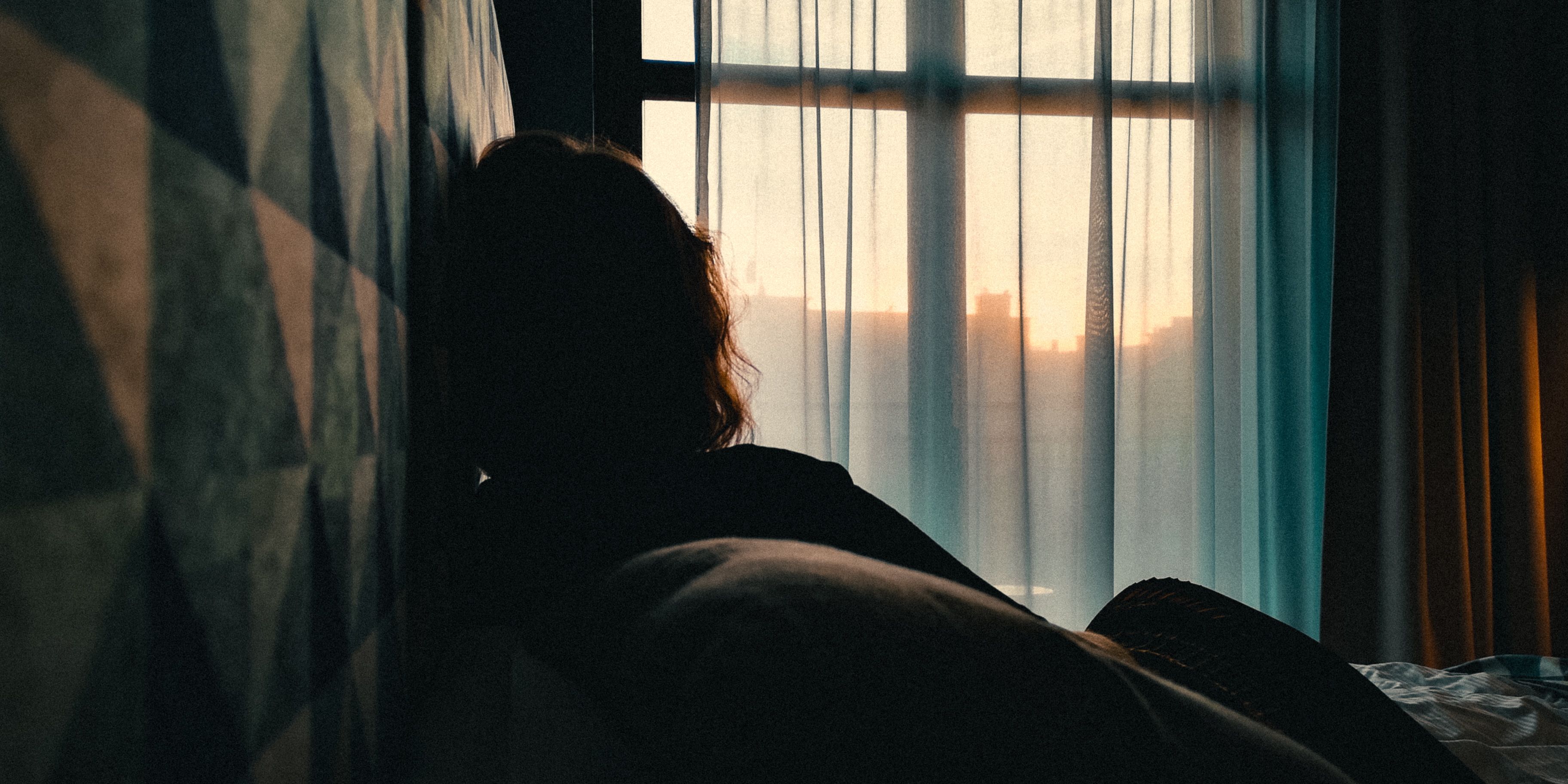 Silhouette of a person looking out a window at sunset with geometric patterned wall. Photo with moody lighting.