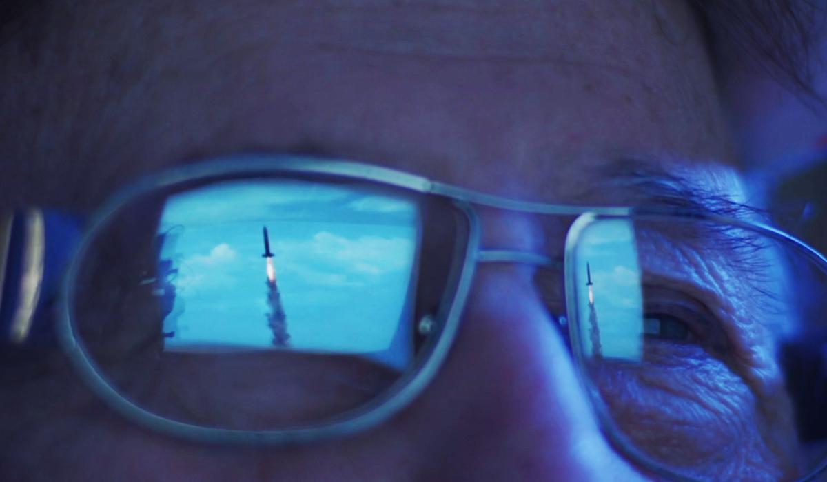 Close-up of a person’s glasses showing the reflection of a rocket launch on a screen, with blue and grey colours dominant.