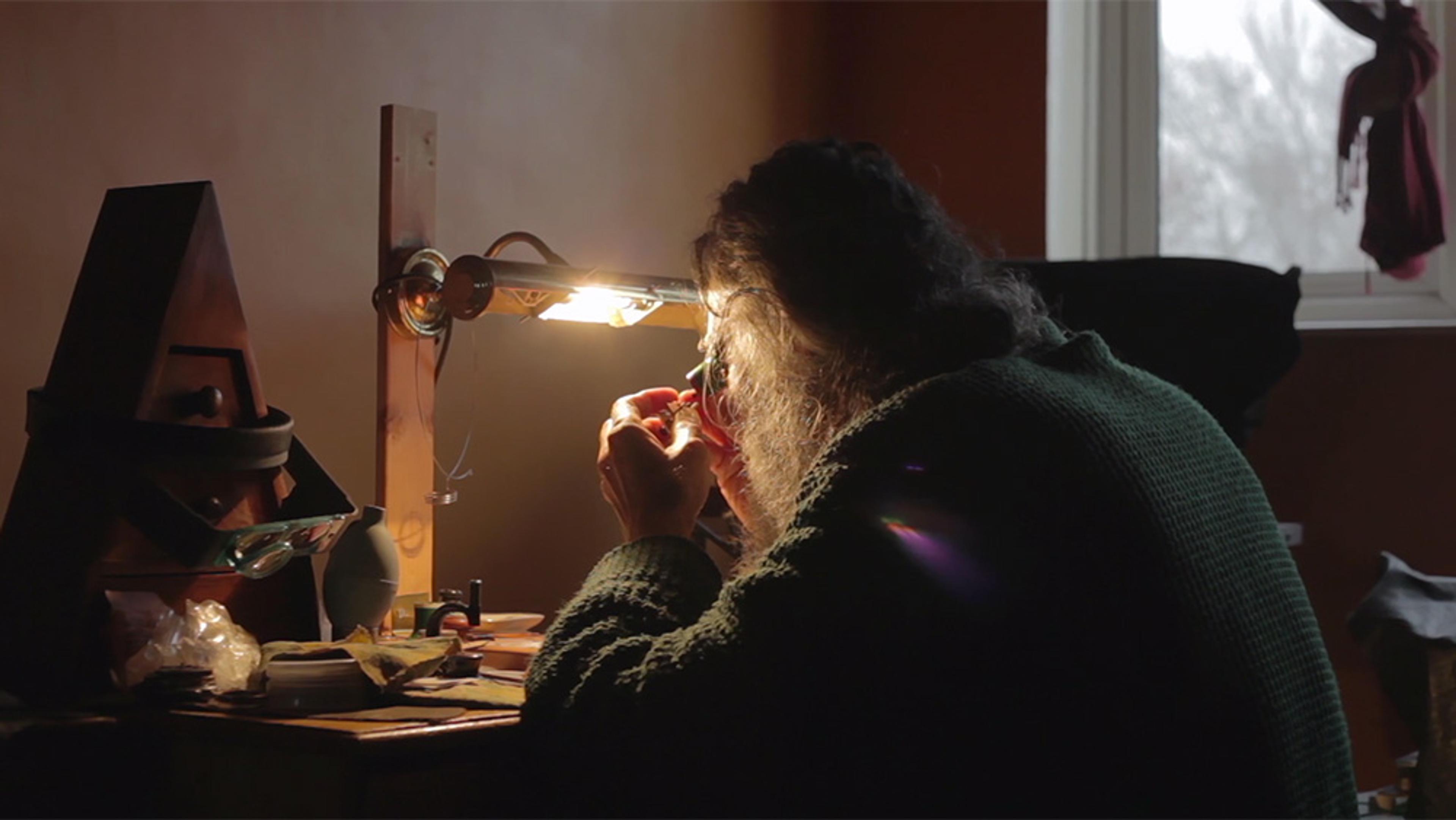 A person focusing intently while working on something small at a cluttered desk lit by a lamp, with a window in the background.