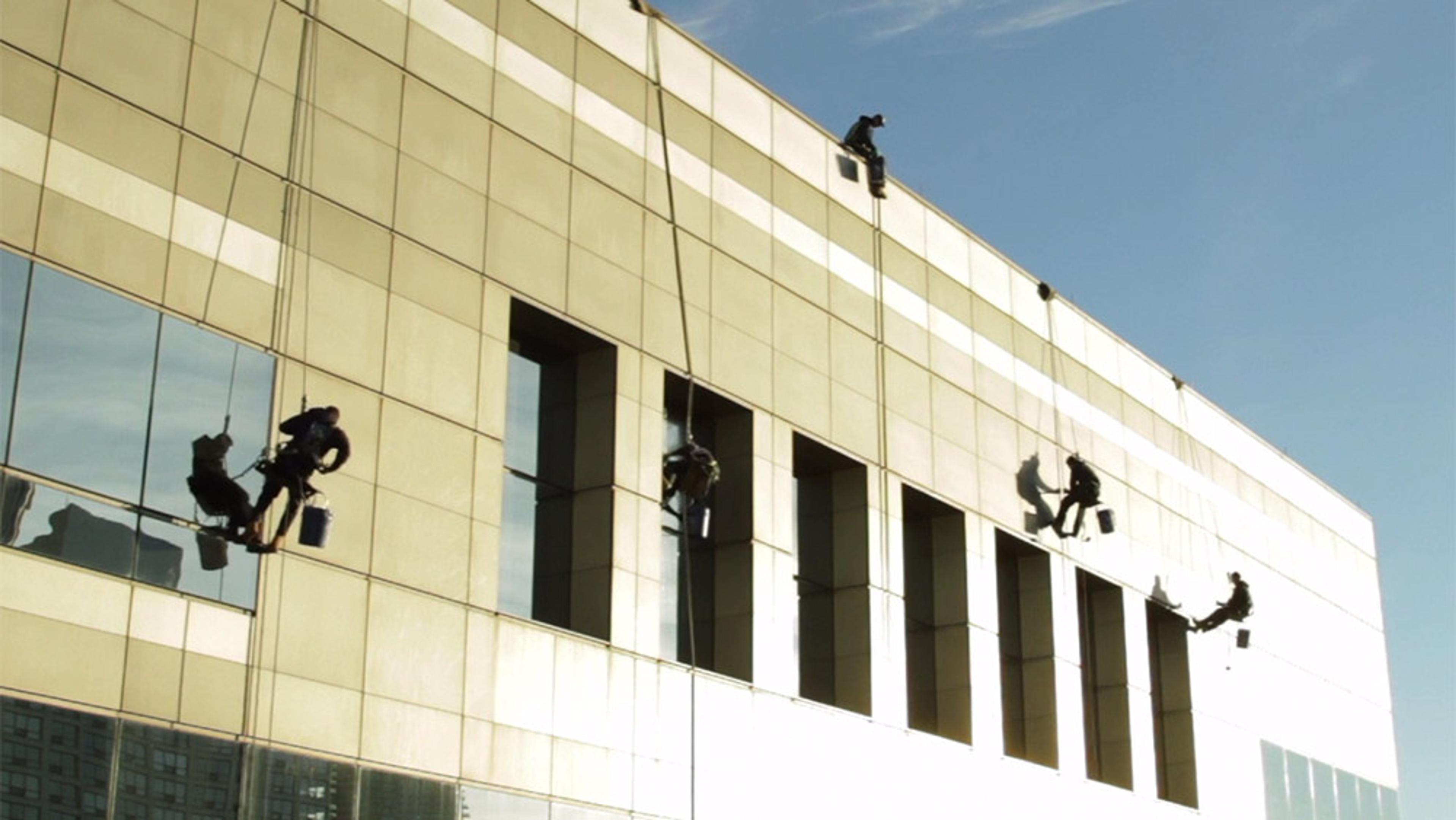 Five window cleaners on ropes cleaning the exterior windows of a modern high-rise building under a clear blue sky.
