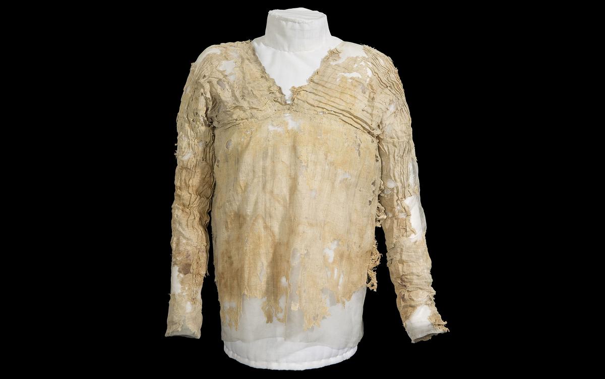 Ancient, tattered linen shirt displayed on a white mannequin against a black background, showing signs of wear and ageing.