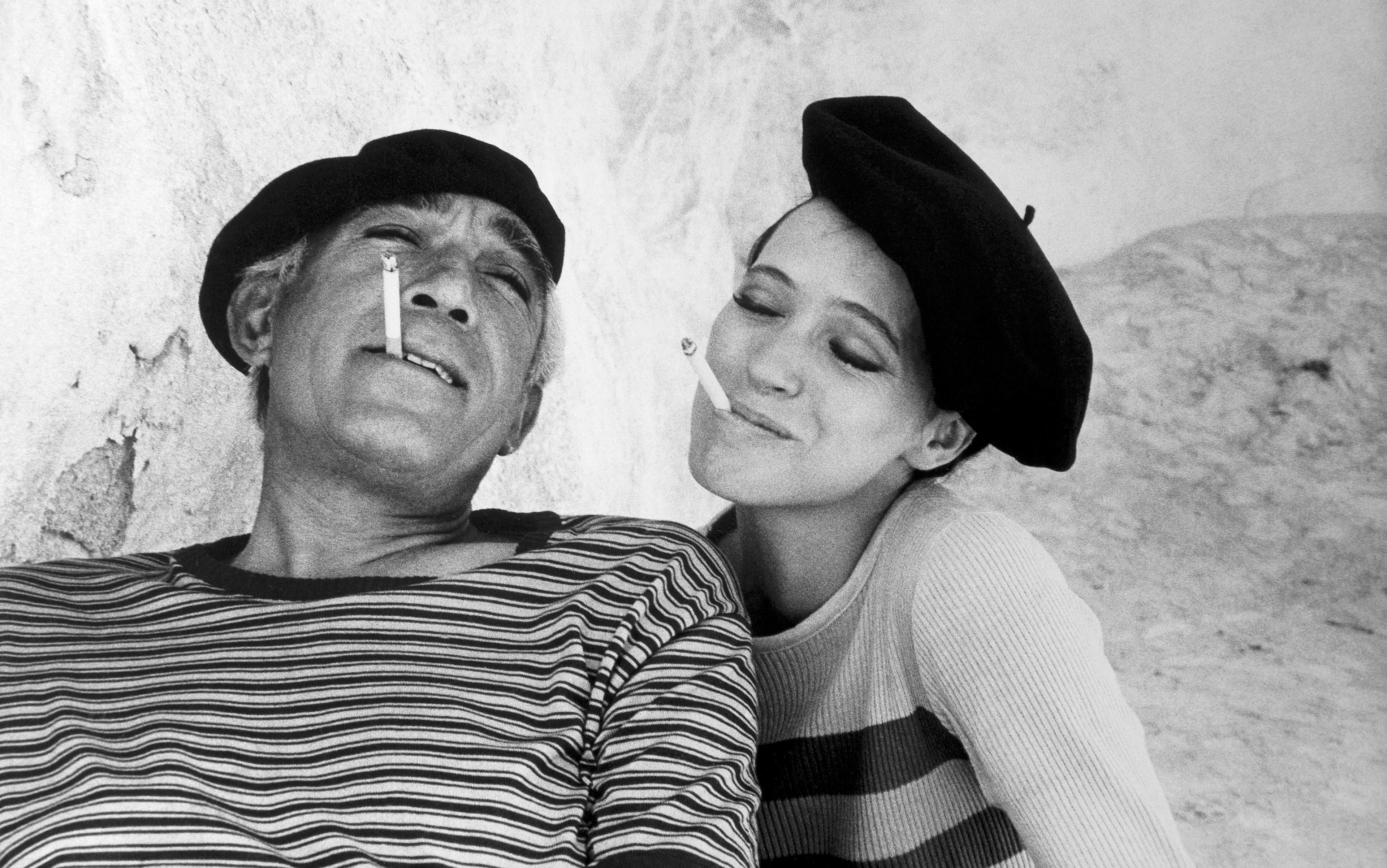 Black and white photo of a man and woman wearing berets, both smiling with cigarettes in their mouths against a textured wall.