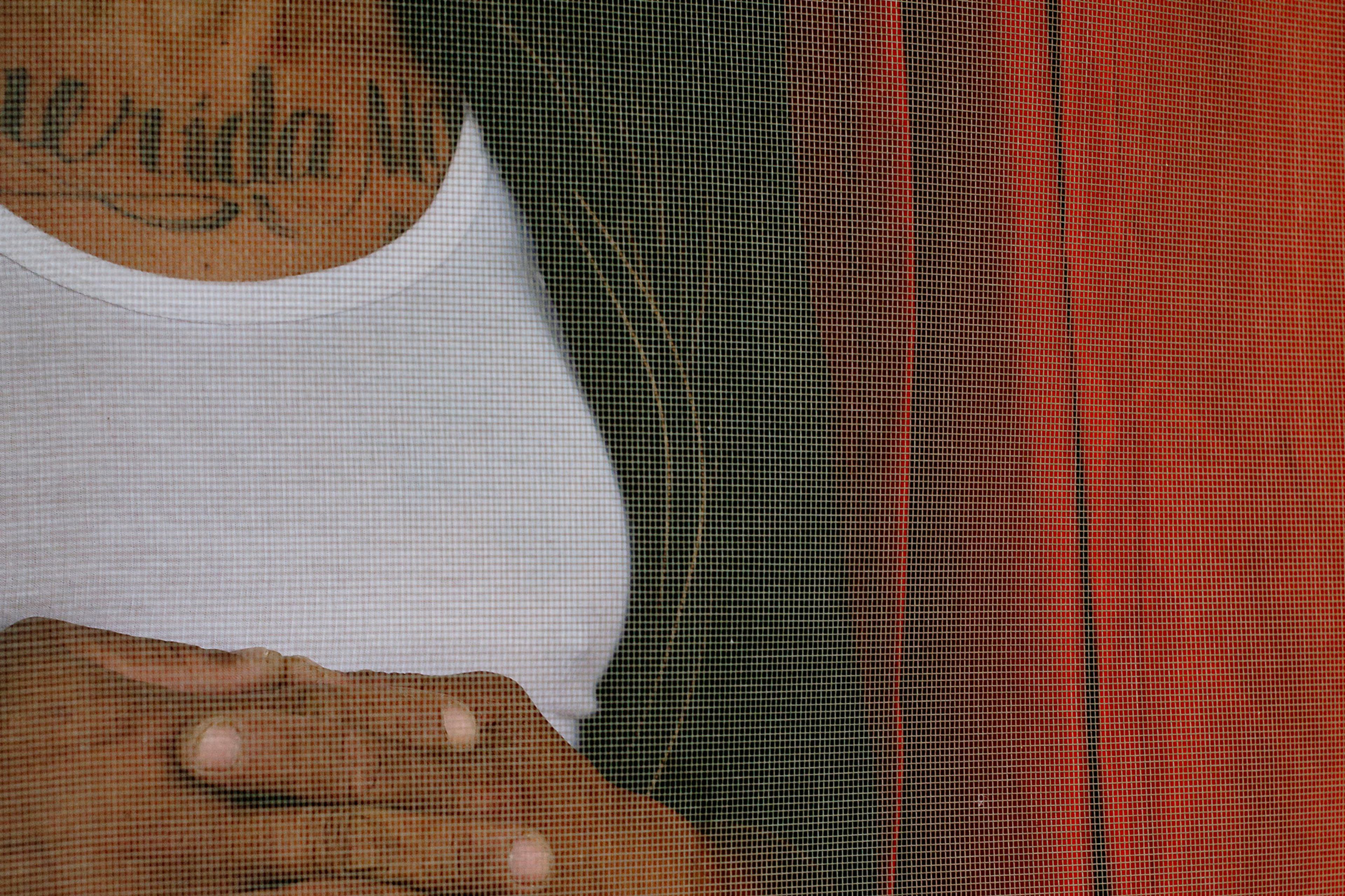 Photo of a person in a white vest and jacket with folded hands and a tattoo, viewed through a mesh screen.