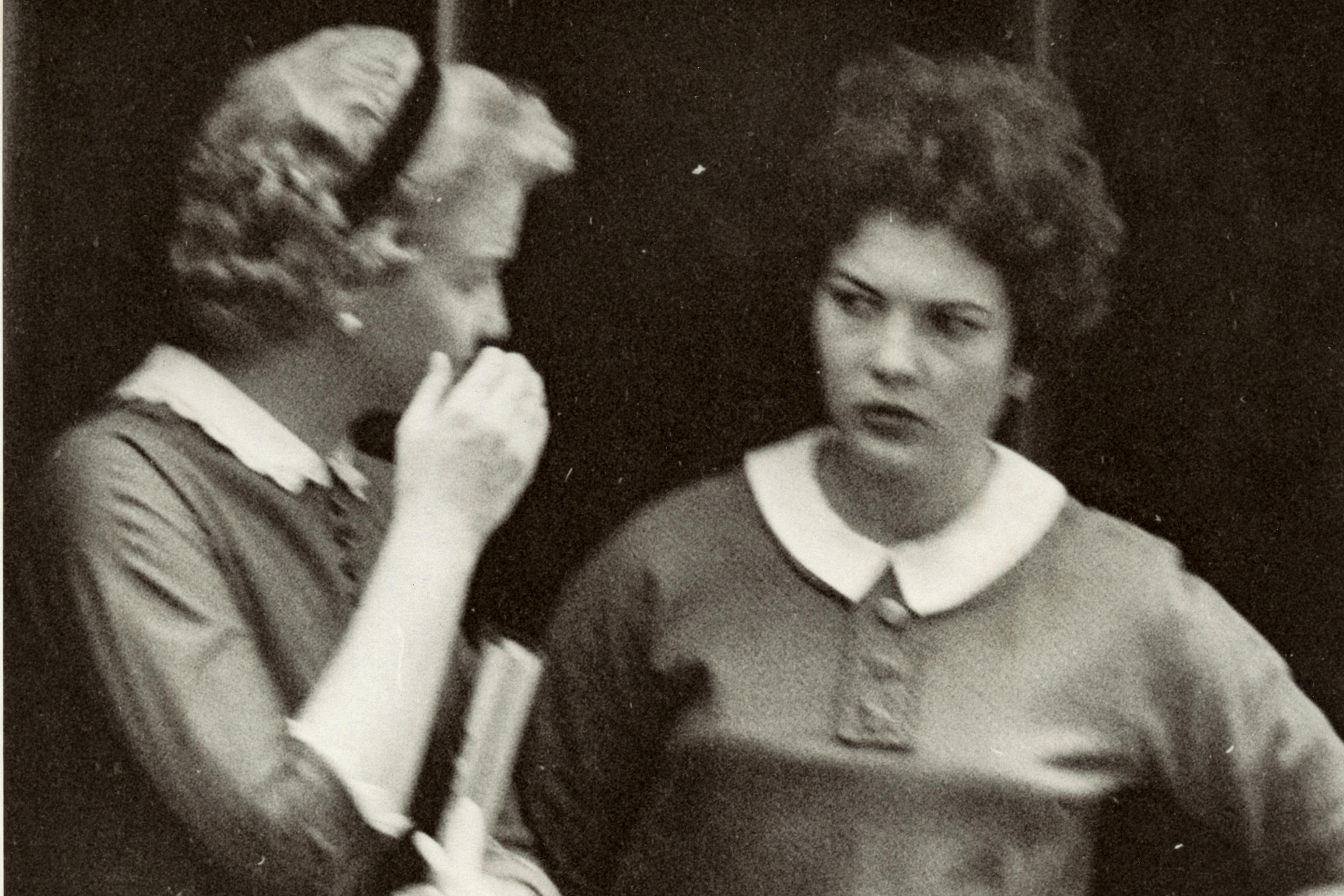 Black and white photo of two women from the 1950s, one whispering to the other.