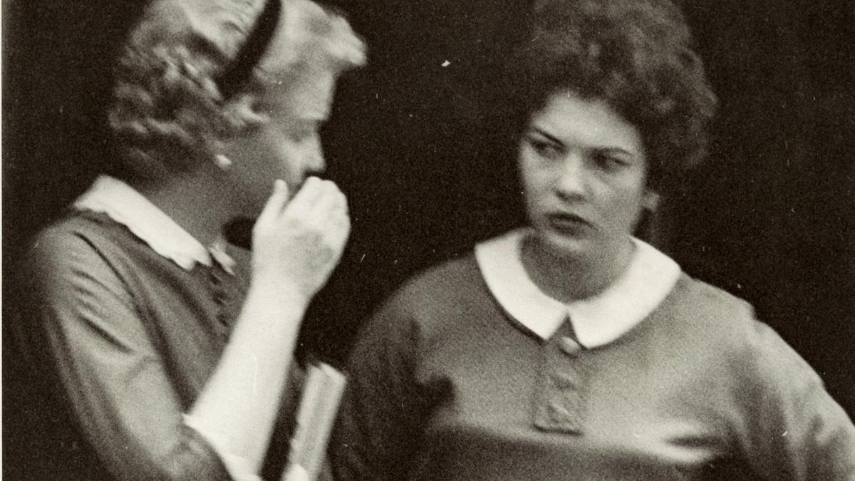 Black and white photo of two women from the 1950s, one whispering to the other.