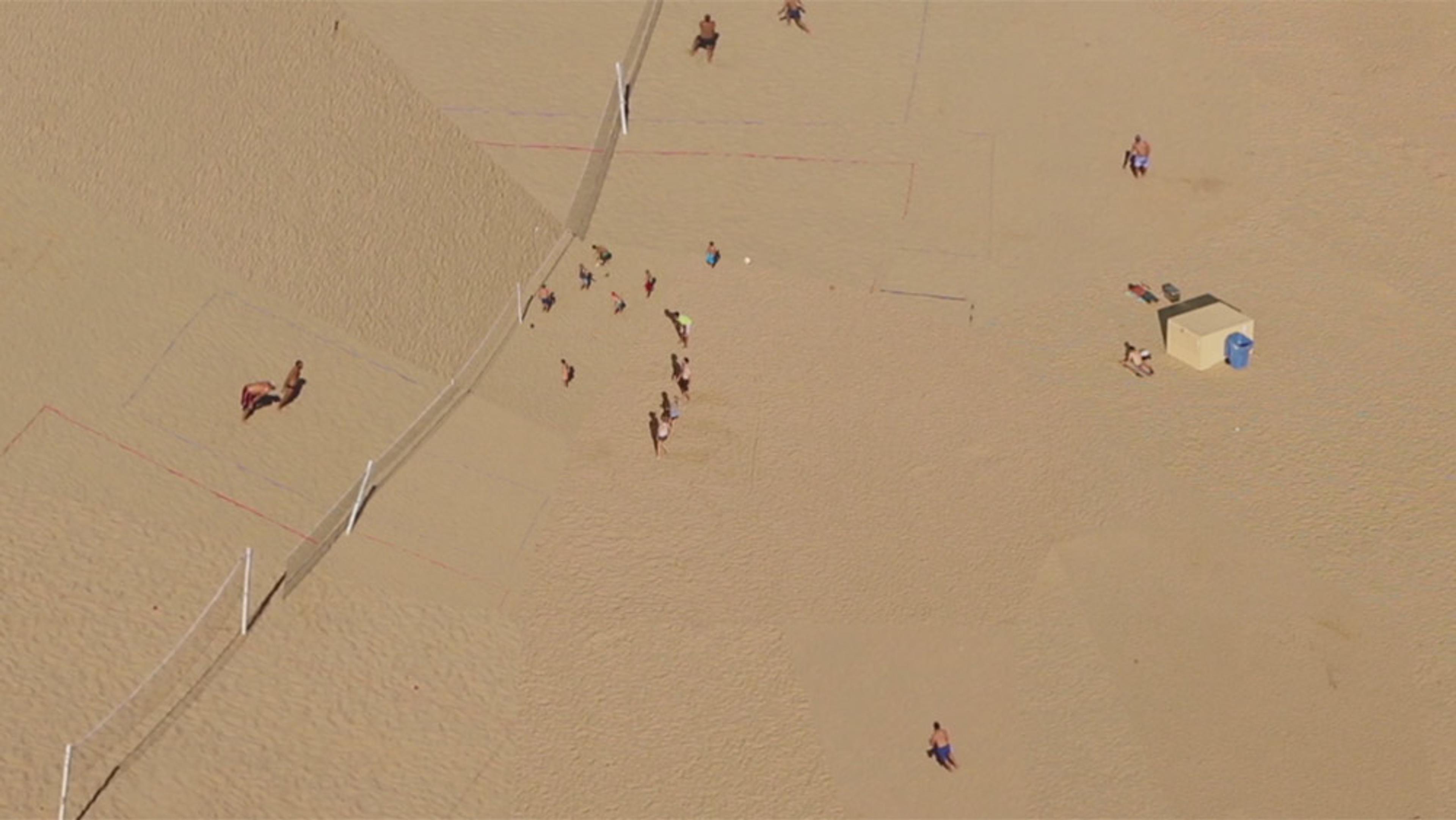 Aerial photo of a sandy beach with people playing volleyball, a few sunbathers and a small shelter in the background.