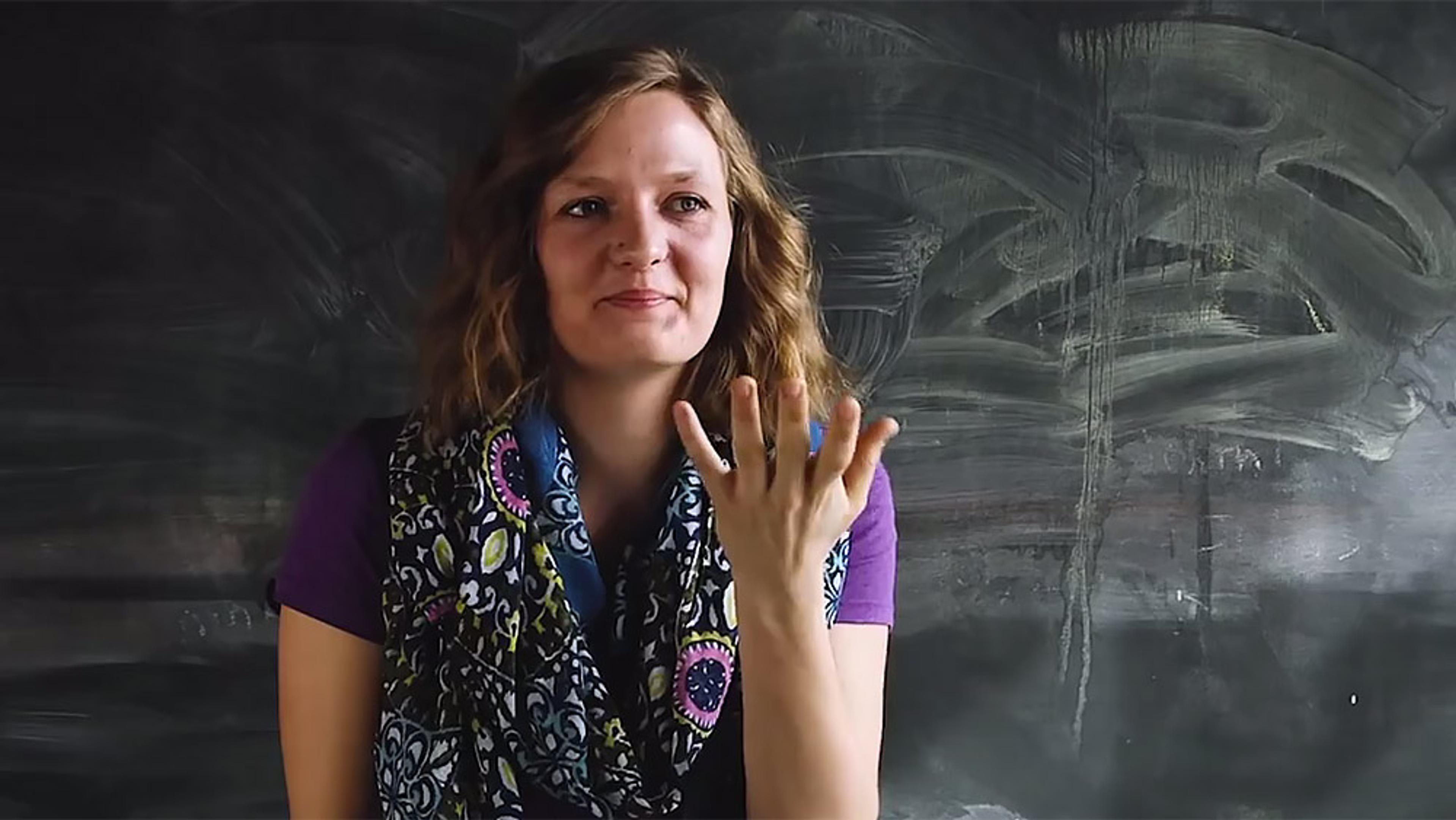A woman with wavy hair in front of a chalkboard, gesturing with her hand and smiling slightly.