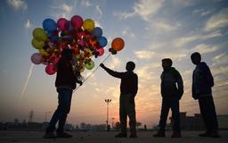 Three silhouetted children at sunset, with one child buying colourful balloons from a seller in an open area with a cloudy sky.