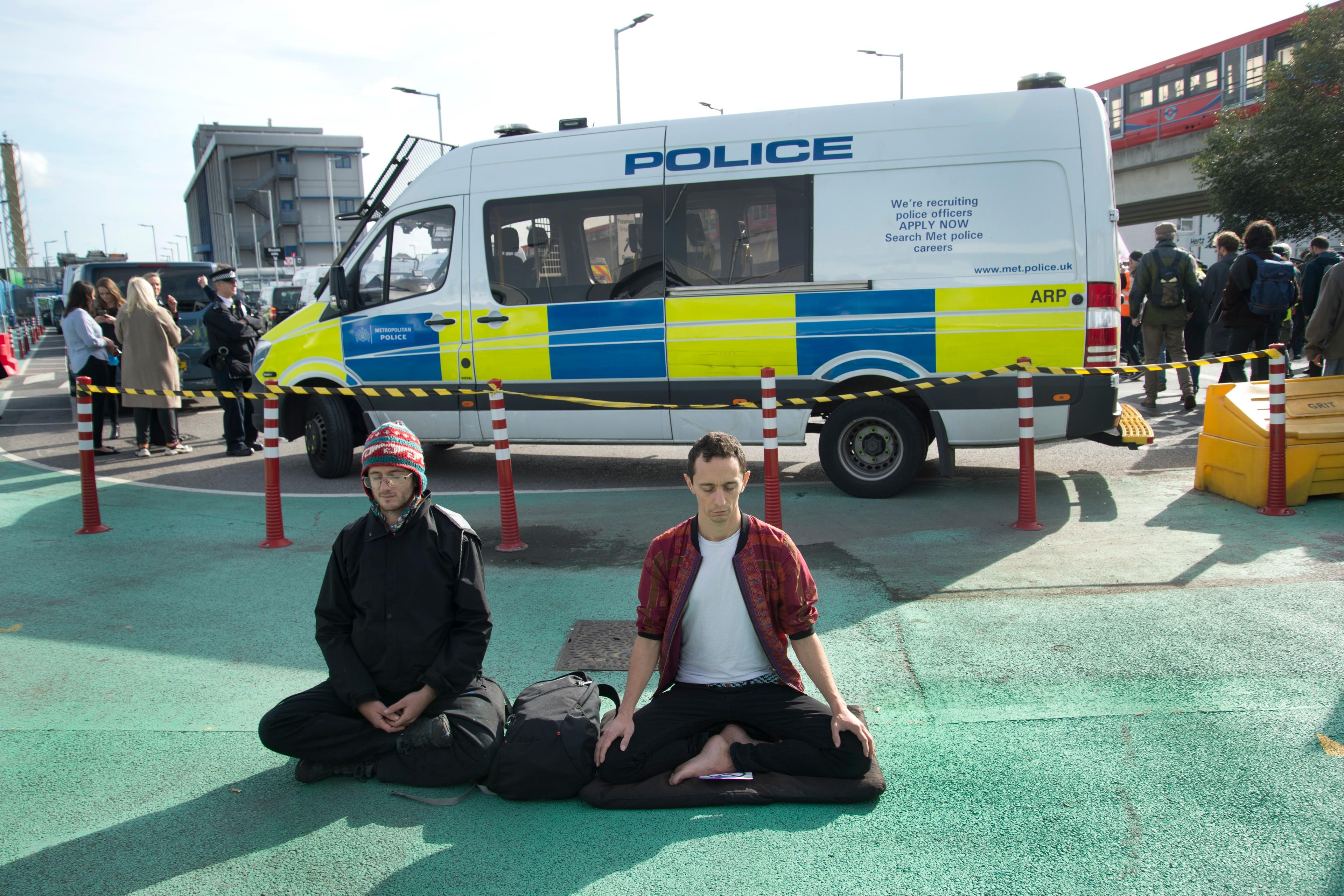 Photo of two people meditating on a road in front of a police van in an urban setting with a few bystanders.