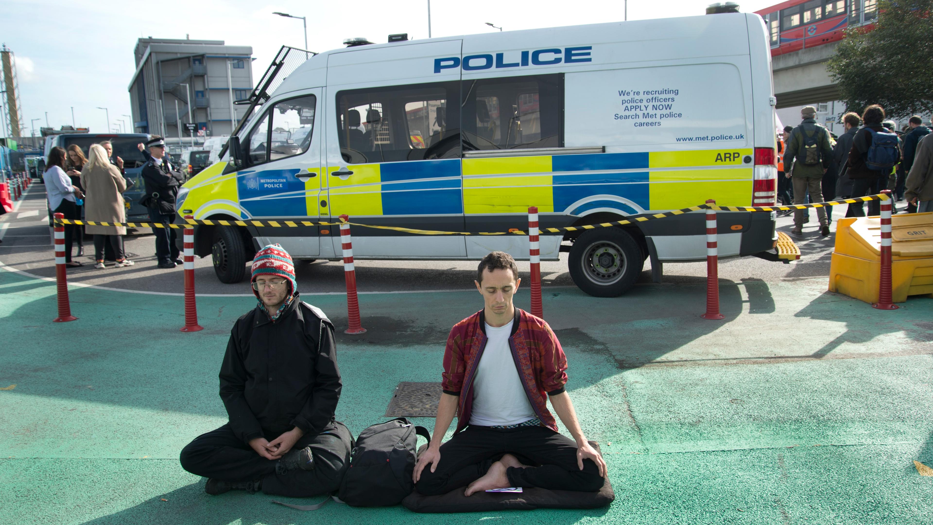 Photo of two people meditating on a road in front of a police van in an urban setting with a few bystanders.