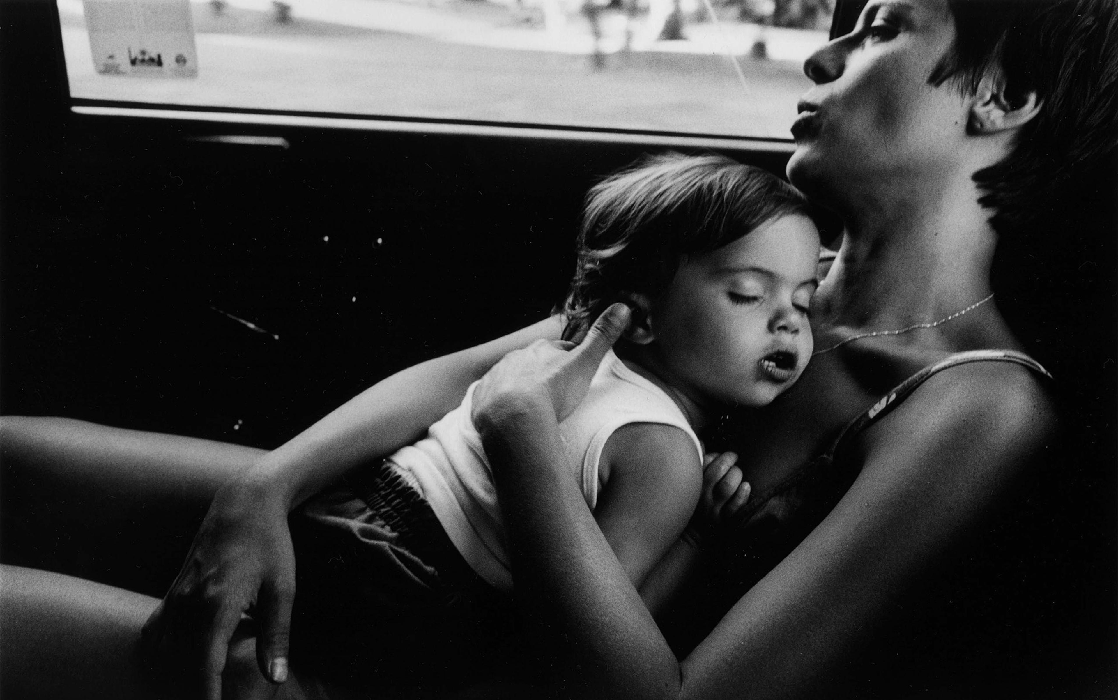 Black and white photo of a woman and a sleeping child in her arms in a moving car, taken from the side showing their upper bodies.